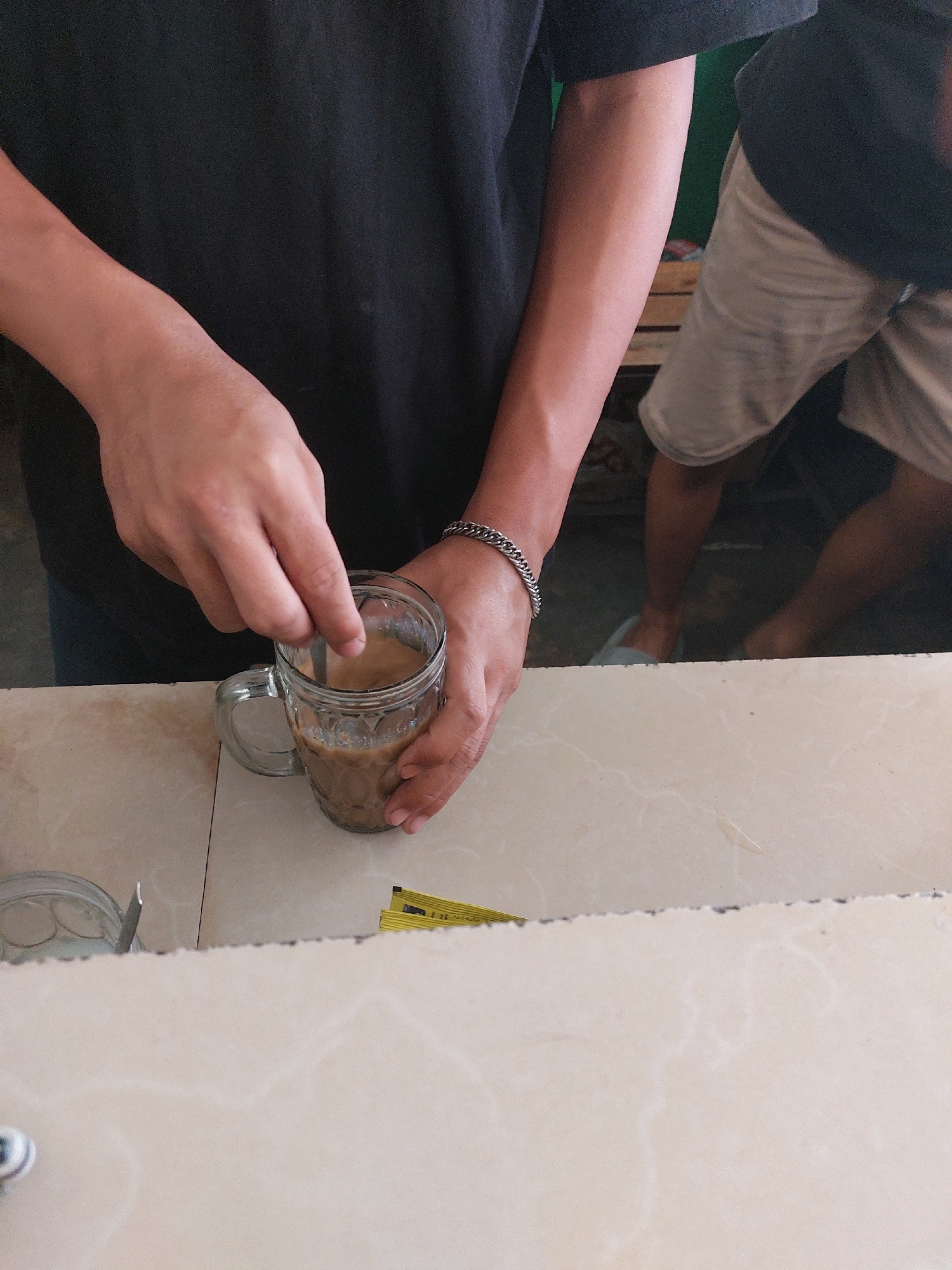 A person's hands stir a beverage in a clear glass mug on a light-colored countertop. The mug contains a light brown liquid, suggesting coffee with milk or creamer. The person wears a dark t-shirt and a silver chain bracelet on their left wrist. In the background, another person wearing beige shorts and a dark t-shirt is visible, along with what appears to be wooden furniture. The setting is likely a cafe or eatery in Sumuragung, Indonesia, given the presence of what looks like packaged sweetener packets on the countertop near the mug. The countertop has a marble-like pattern.