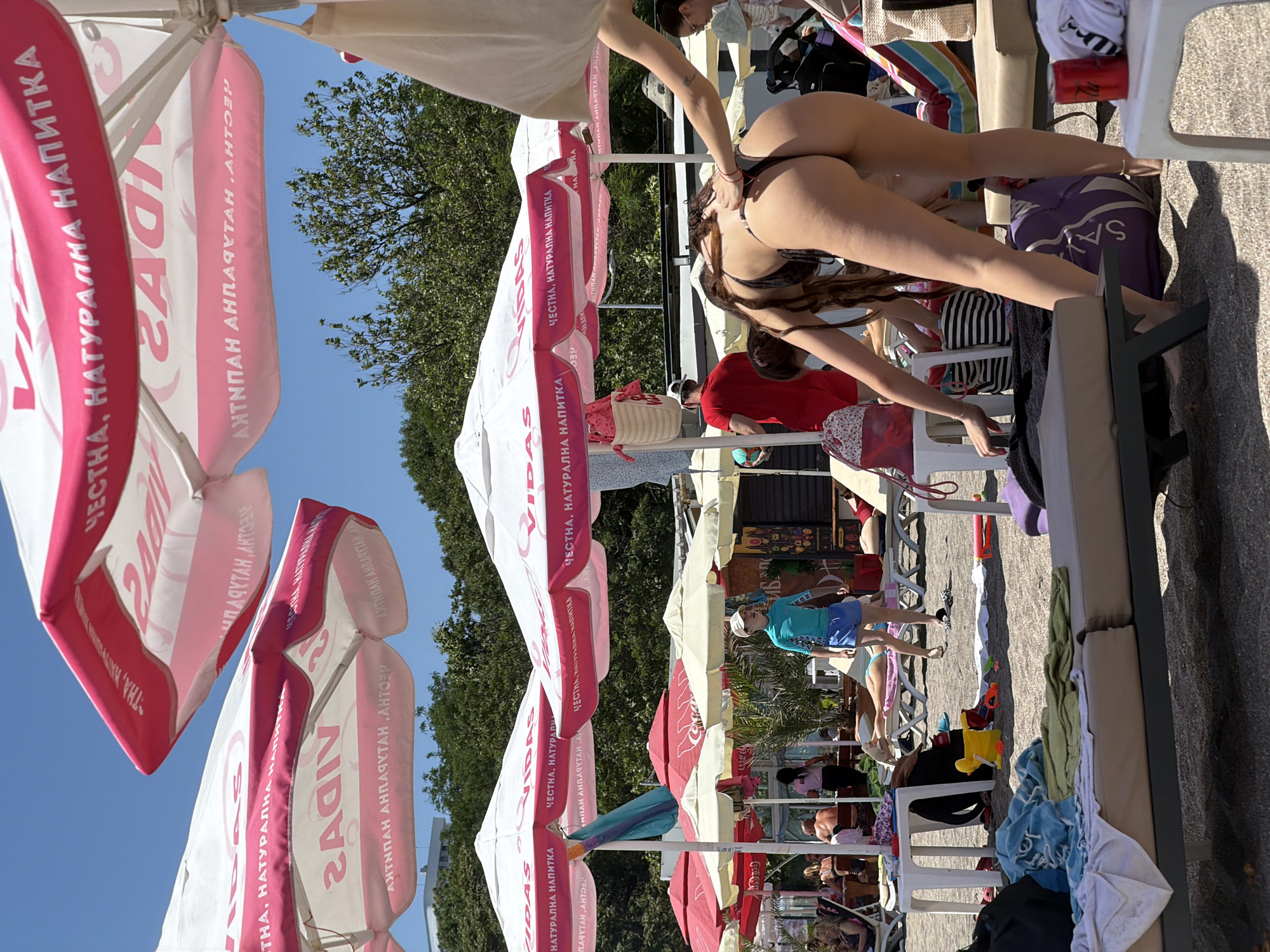 On a sunny day at Burgas' South Beach, Bulgaria, people relax on lounge chairs under large pink and white umbrellas. The umbrellas display text, possibly a brand name, in Cyrillic script. A young woman in a black bikini is bent over, adjusting her swimwear, with her back to the camera. Another person's hand is visible on her back. In the background, a child in a blue swimsuit walks on the sand. Beachgoers are scattered across the scene, some lying down, others walking. Various beach accessories, including towels, bags, and toys, are visible on the sand and on tables. The environment is a typical busy beach scene with sand, umbrellas, and trees in the distance.