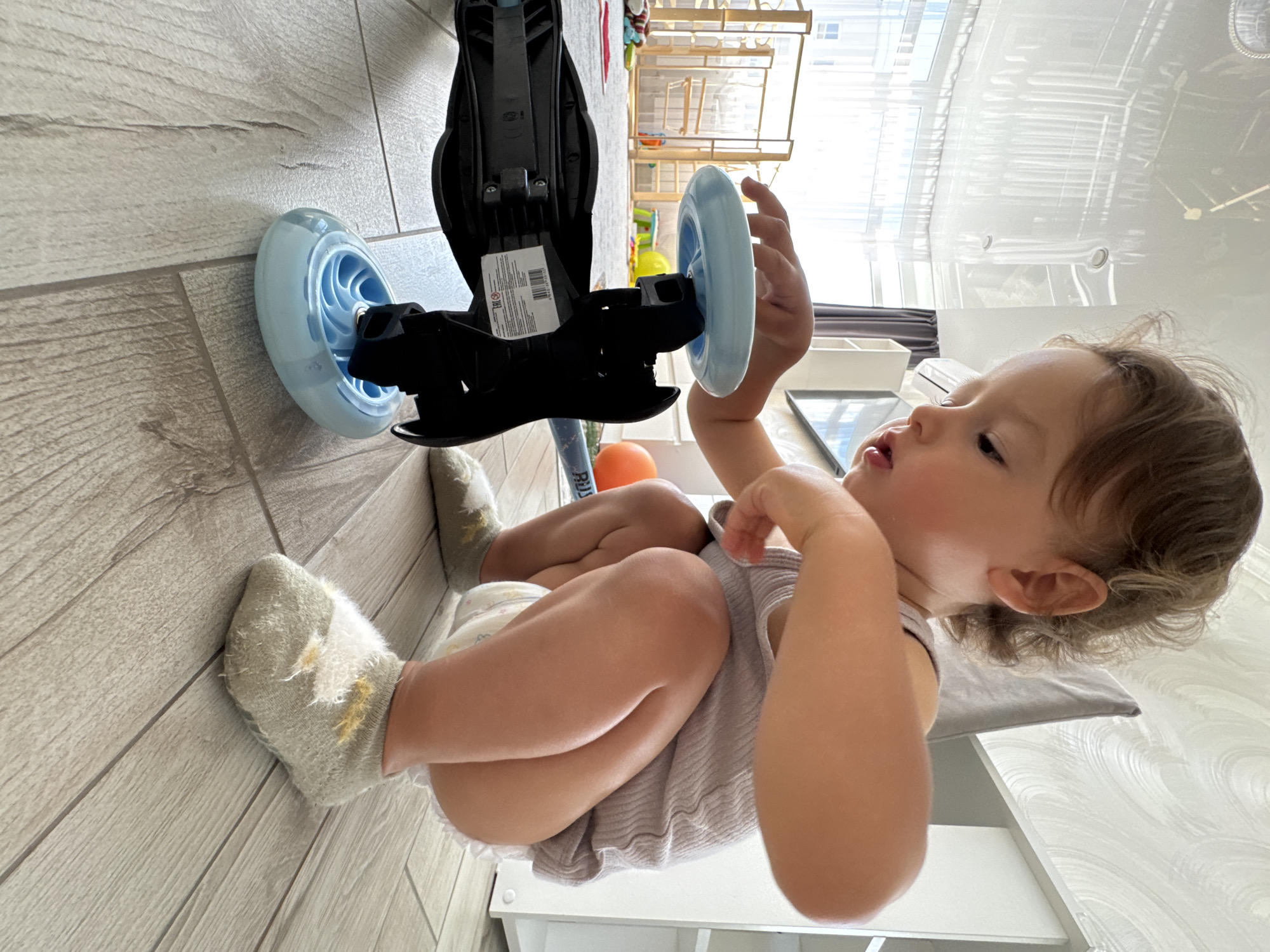 A toddler is crouched on a wooden floor, focused on a blue scooter. The child, wearing a light grey tank top and a diaper, has light brown curly hair. Their feet are covered in fuzzy grey socks with yellow accents. The scooter is black and blue with three wheels. In the background, a large window reveals a glimpse of what appears to be a bright, sunlit room with a playpen and an orange ball. The ceiling features recessed lighting and a reflective surface. The overall scene suggests an indoor play area, potentially in a residential setting. The image does not provide any specific information to indicate the location is in Sochi, Russia.