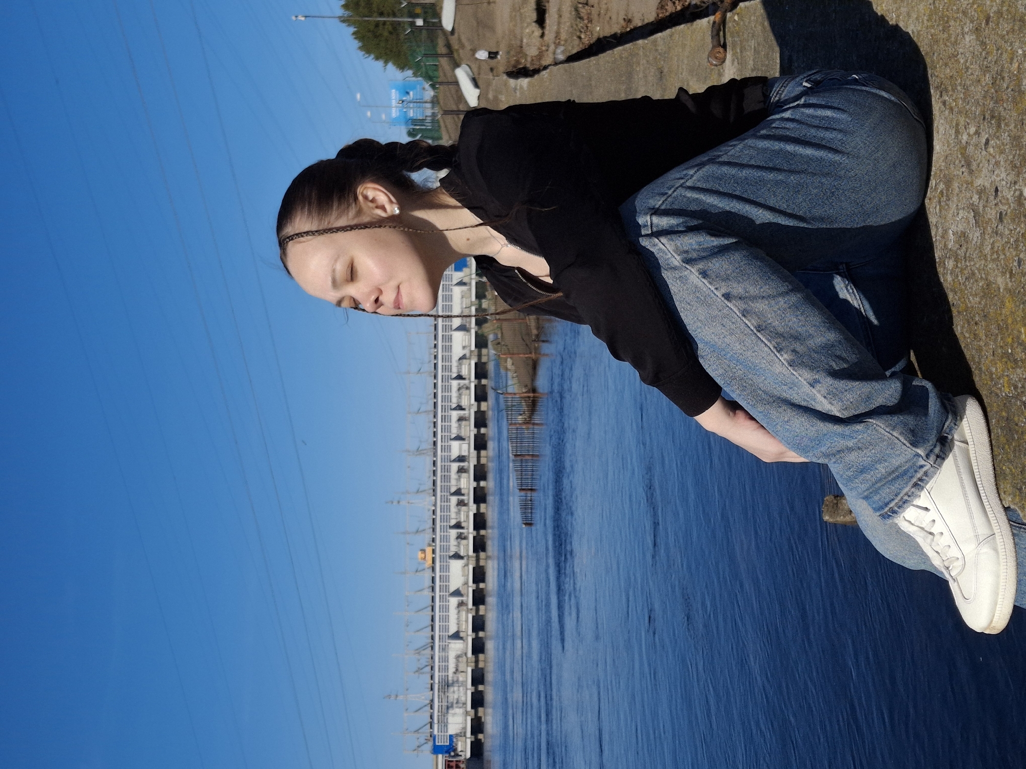 A young woman is prominently featured, leaning or sitting against a rough, sun-drenched concrete embankment, with strong shadows visible behind her. She has dark hair, styled with thin braids framing her face, and is wearing a black long-sleeved top, blue jeans, and white sneakers. Her eyes are closed, and her head is tilted downwards, suggesting a moment of contemplation, rest, or peacefulness. A silver necklace and a single earring are visible.
The scene is set outdoors by a wide, deep blue body of water, likely a river. Across the water, a significant man-made structure extends horizontally, resembling a large dam or a hydroelectric power station with multiple segments and associated pylons. The sky above is a clear, bright blue, without clouds, indicative of a sunny day, possibly mid-morning or afternoon given the defined shadows. Thin power lines stretch across the sky. The location context of Chaikovsky, Russia, strongly suggests this is the Votkinsk Hydroelectric Power Station on the Kama River. There is no visible text in the image.