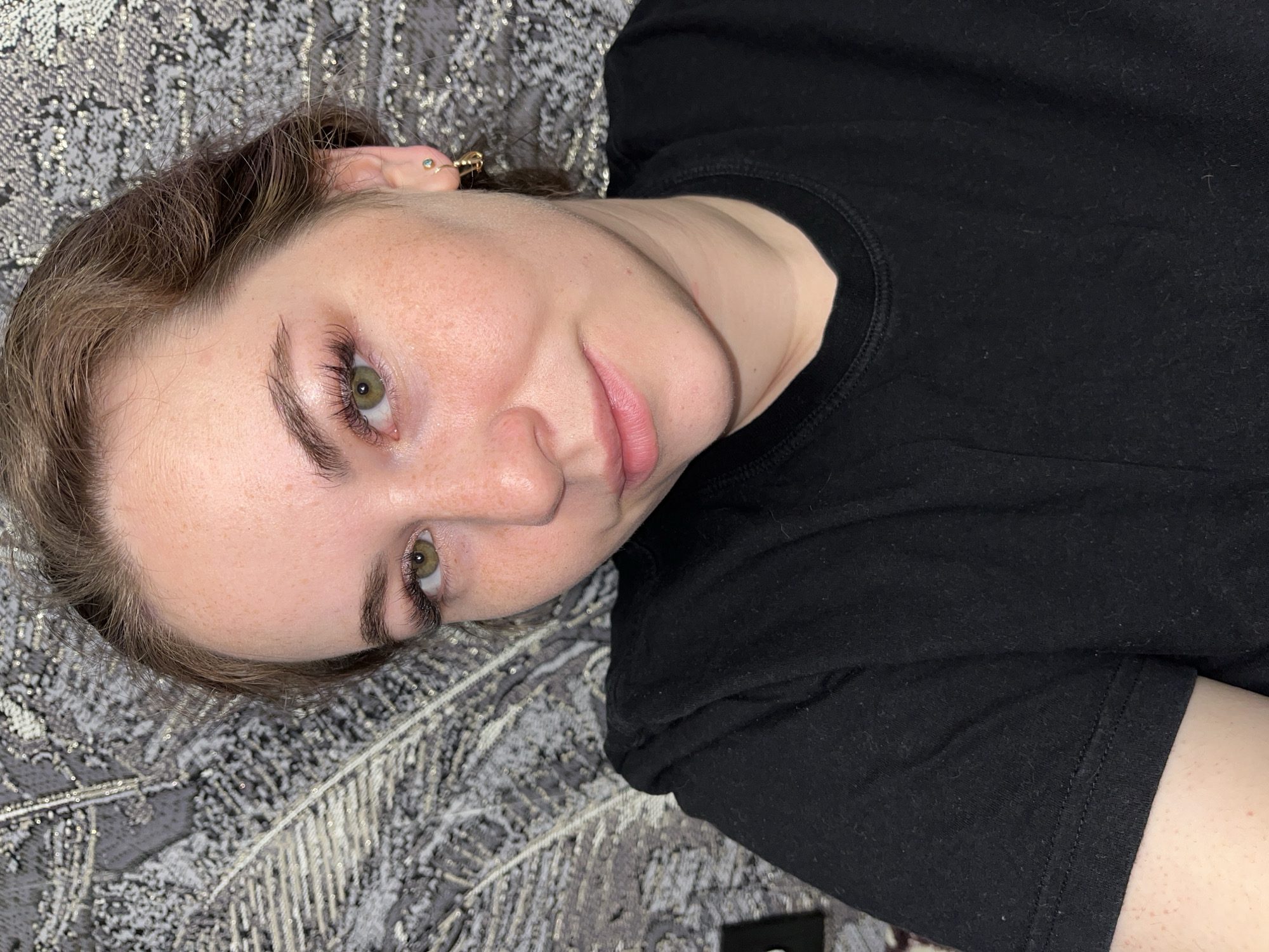 A person with fair skin and light brown hair is lying on a gray patterned surface. The person is wearing a black shirt. They have green eyes and visible freckles on their face. An earring is visible in the left ear. The location in the city of Chaikovsky, Russia is not discernible from this image.