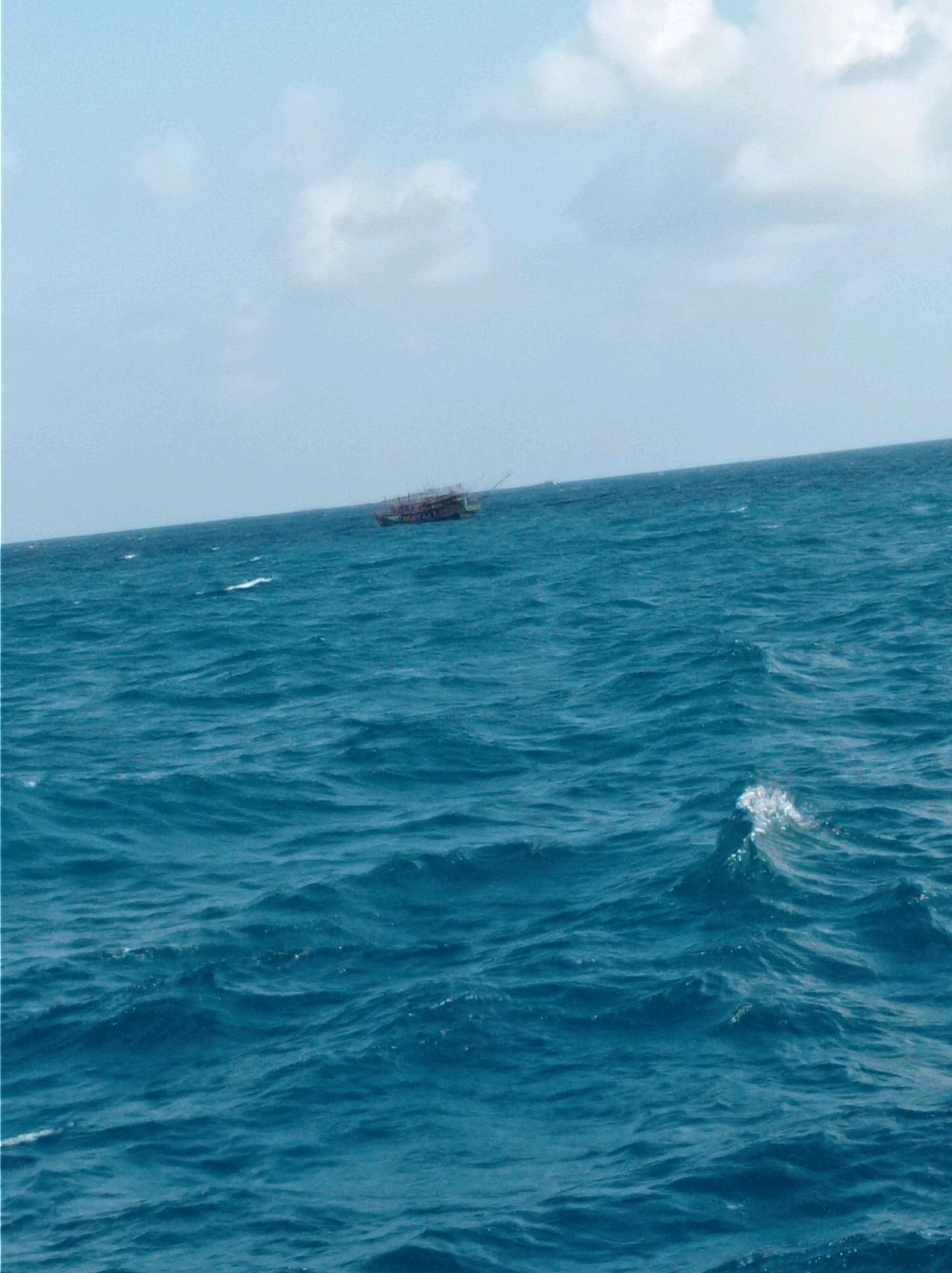 A distant, small, multi-decked fishing vessel, appearing to be a trawler, is visible on a vast expanse of choppy, deep blue ocean. The boat has a cluttered deck with visible structures and netting, suggesting it is actively engaged in fishing. The water is a vibrant turquoise, with whitecaps and ripples indicating moderate waves. The sky above is a pale blue, with scattered, wispy white clouds. The horizon line is clearly defined, separating the sea from the sky. The overall scene depicts a typical day at sea for a fishing operation in a coastal Indonesian location.