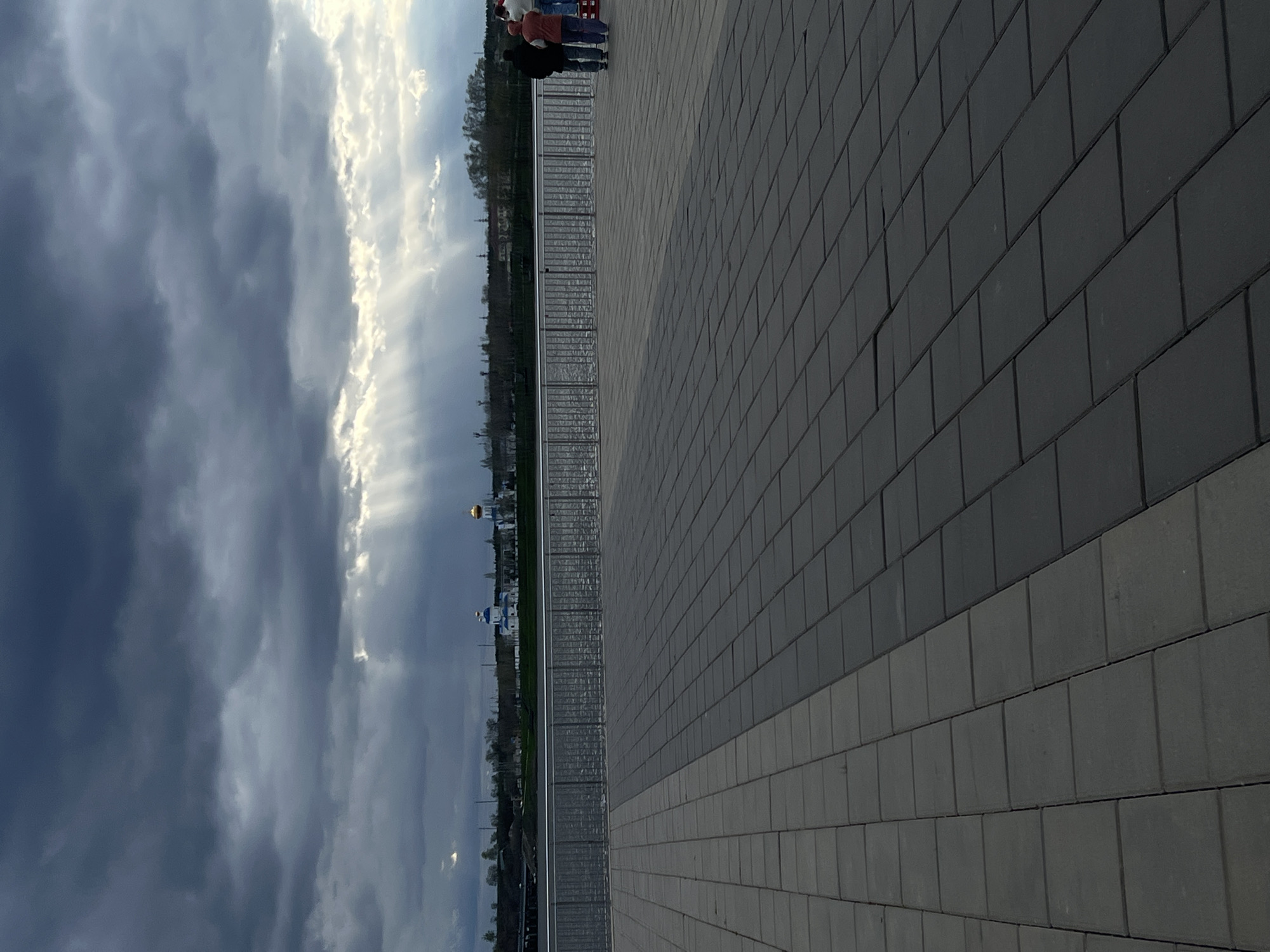 The image depicts a street scene in Syzran, Russia. A paved walkway made of rectangular blocks of varying shades of gray occupies the right half of the frame. A metal railing runs along the edge of the walkway, separating it from a lower level with vegetation. Beyond the railing, there is a view of the cityscape, including a golden dome and a white and blue church, and a dark cloudscape. Two individuals are standing near the railing.