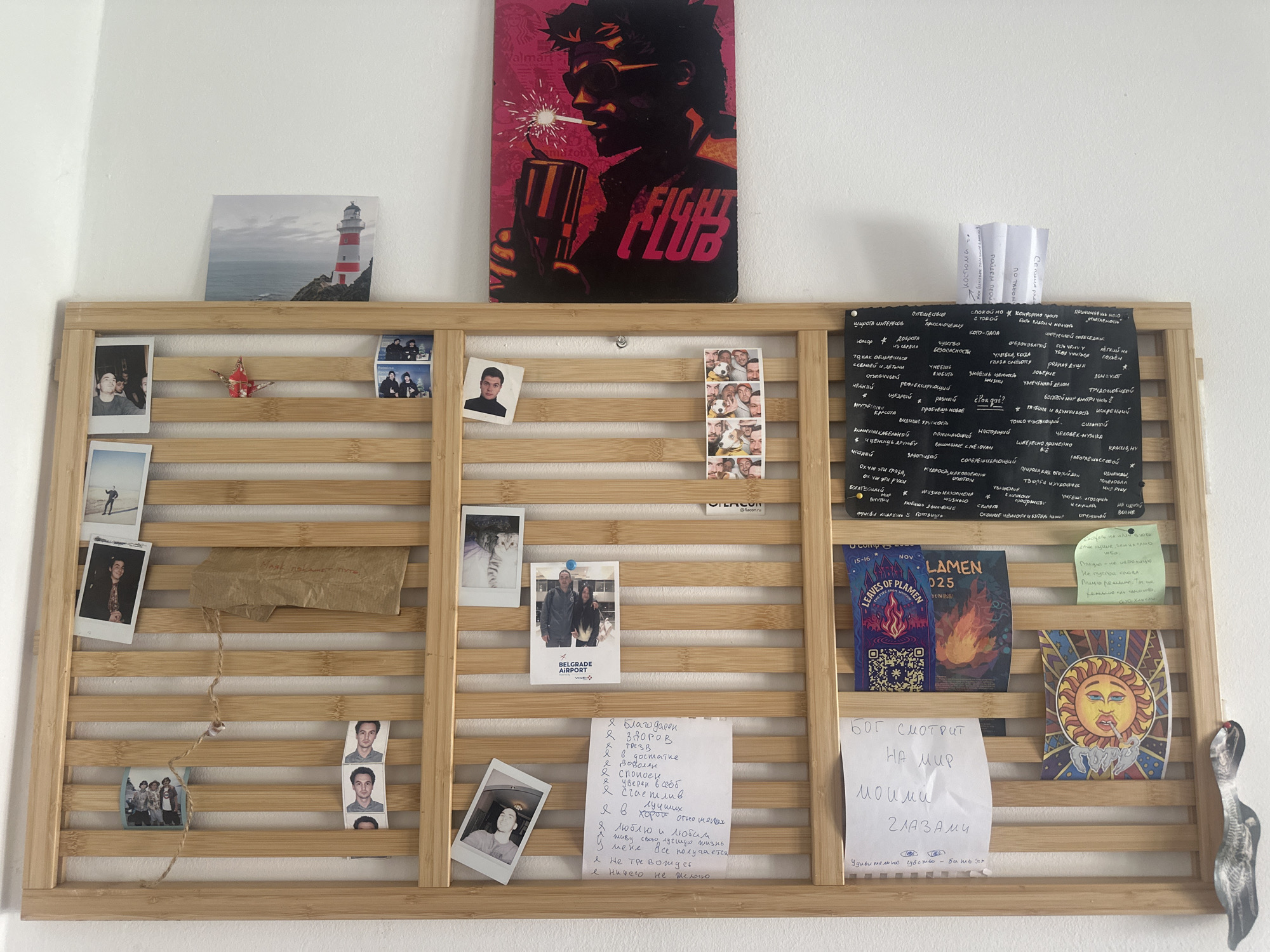 The image shows a wooden slatted wall organizer filled with various items. The organizer is mounted on a white wall. On the top shelf of the organizer, there is a framed picture of a lighthouse and a red origami crane. To the right of these, there are several small photos.
The organizer is filled with a mix of personal items, including photographs, notes, and flyers. In the center section, there are several more photos, including some polaroid pictures and a strip of photo booth pictures. There is also a small piece of crumpled brown paper with writing on it.
The bottom section of the organizer contains more notes and flyers. One flyer has the title "LEAVES OF PLAMEN 025" with flames. Another flyer is a colorful depiction of the sun with a face. There are also handwritten notes on white paper.
On the far left, there are several polaroid photos attached to the organizer, showing people. On the far right, a black notice board is attached to the organizer, covered in handwritten text.
There are no visible clues or elements within the image that suggest the location is Budva, Montenegro. The scene appears to be an indoor personal display area.