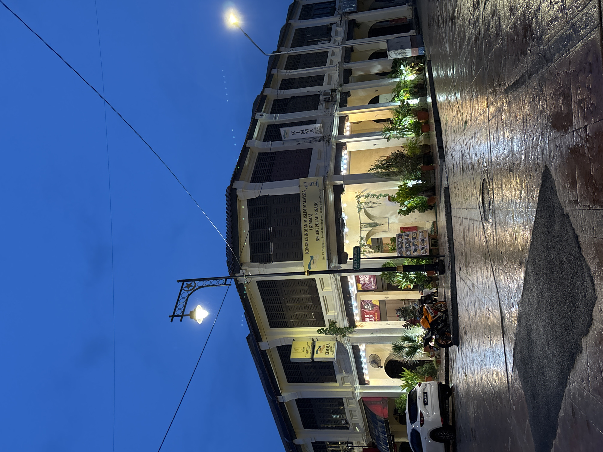 The image captures a street scene in George Town, Malaysia, at dusk, with the sky a deep blue and streetlights illuminating the wet pavement, reflecting the warm glow of the building's interior lights and street lamps. The architecture is colonial, characterized by white buildings with dark wooden shutters. In the foreground, a white SUV with license plate "CEM 889" is parked on the left. To its right, an orange and black motorcycle is parked. A man in a white shirt and dark pants is standing near the motorcycle. A street sign indicates "LEBUH CARNARVON". The building displays signs for "KIMMA PULAU PINANG" and "KONGRES INDIAN MUSLIM MALAYSIA". The wet street suggests recent rain. Plants in pots are visible along the building's facade. The overall impression is of a quiet, atmospheric evening in a historic urban setting.