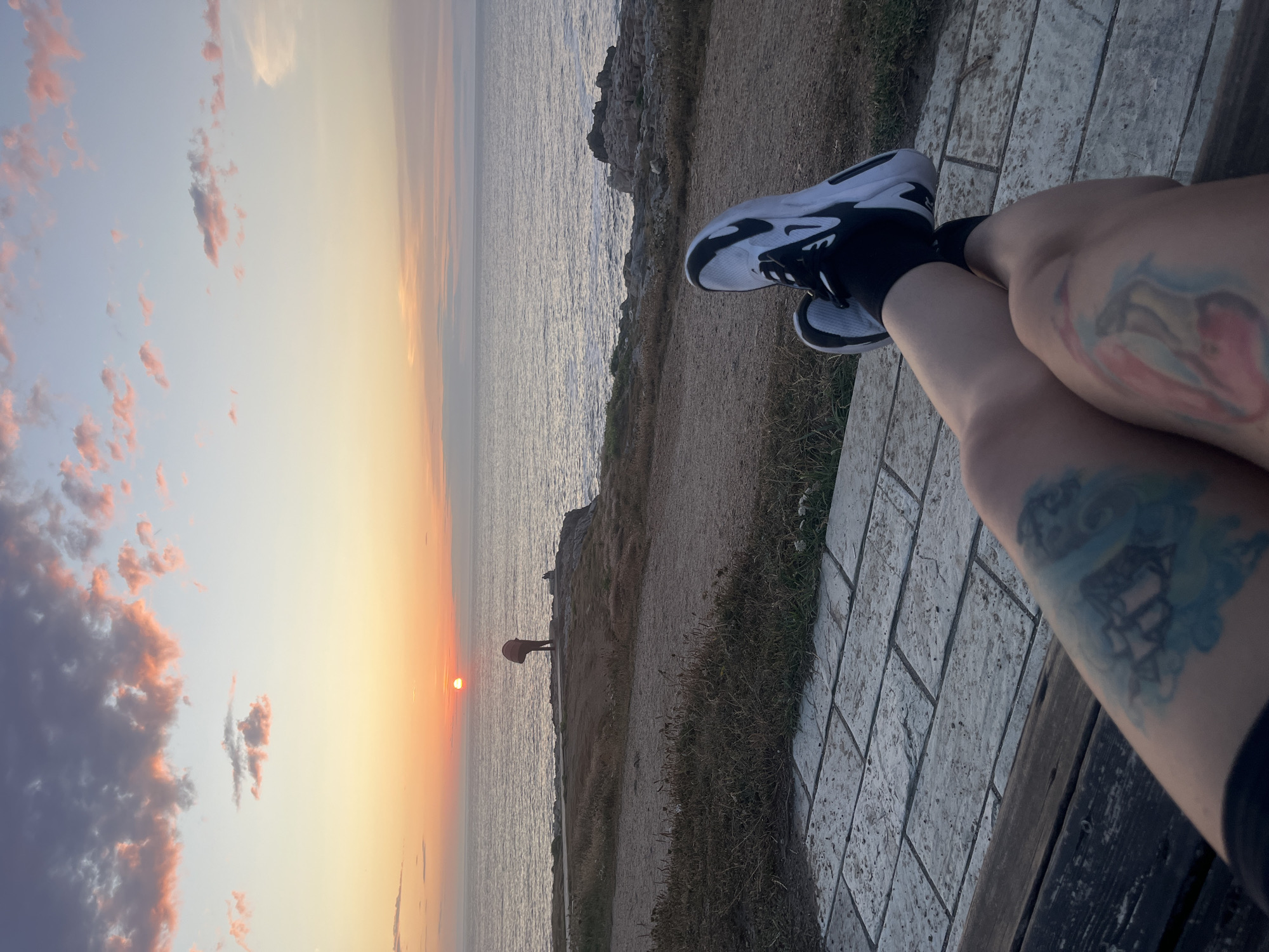 The image shows a view of the Atlantic Ocean at sunset, with the sun low on the horizon and casting a warm glow across the sky and water. The scene is captured from a seated perspective, with a person's legs and feet, adorned with tattoos, in the foreground. The person is wearing athletic shoes and socks. In the middle ground, a path leads to a cliff overlooking the ocean, where a lone figure stands with an umbrella. The coast is rocky and lined with dry grass. The sky is filled with soft clouds, tinged with pink and orange from the setting sun. This scene is typical of the coastline of A Coruña, Spain, known for its dramatic sunsets over the Atlantic.