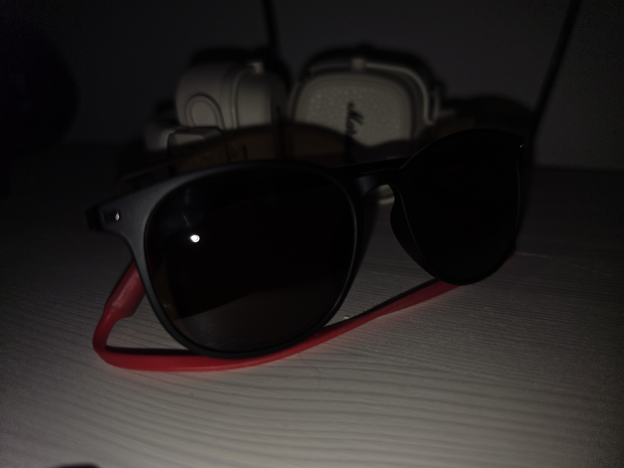 A pair of black-framed sunglasses with dark lenses rests on a light-colored, horizontally-grained surface, possibly a table. A bright red cord is attached to the arms of the sunglasses and loops underneath them. In the blurred background, two white-cased headphones with visible logos are stacked. The lighting is dim, creating shadows and highlighting the reflective lenses of the sunglasses. There are no people or any specific indications of the location in Strelna, Russia.
