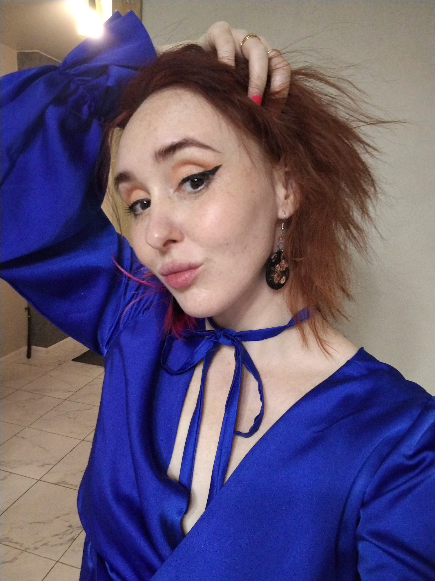 A fair-skinned woman with red hair is posing for a selfie. She is wearing a blue silk dress with a V-neck and a ribbon tie. The dress has large, billowy sleeves. The woman is wearing earrings and has gold rings on her fingers. She has red nail polish on. Her makeup includes winged eyeliner and peachy eyeshadow. She is holding her hair with one hand and making a kissy face at the camera. The background is a neutral-colored wall and a tiled floor. It is unlikely the photo was taken in Izhevsk, Russia, as there is no indication of the location in the image.