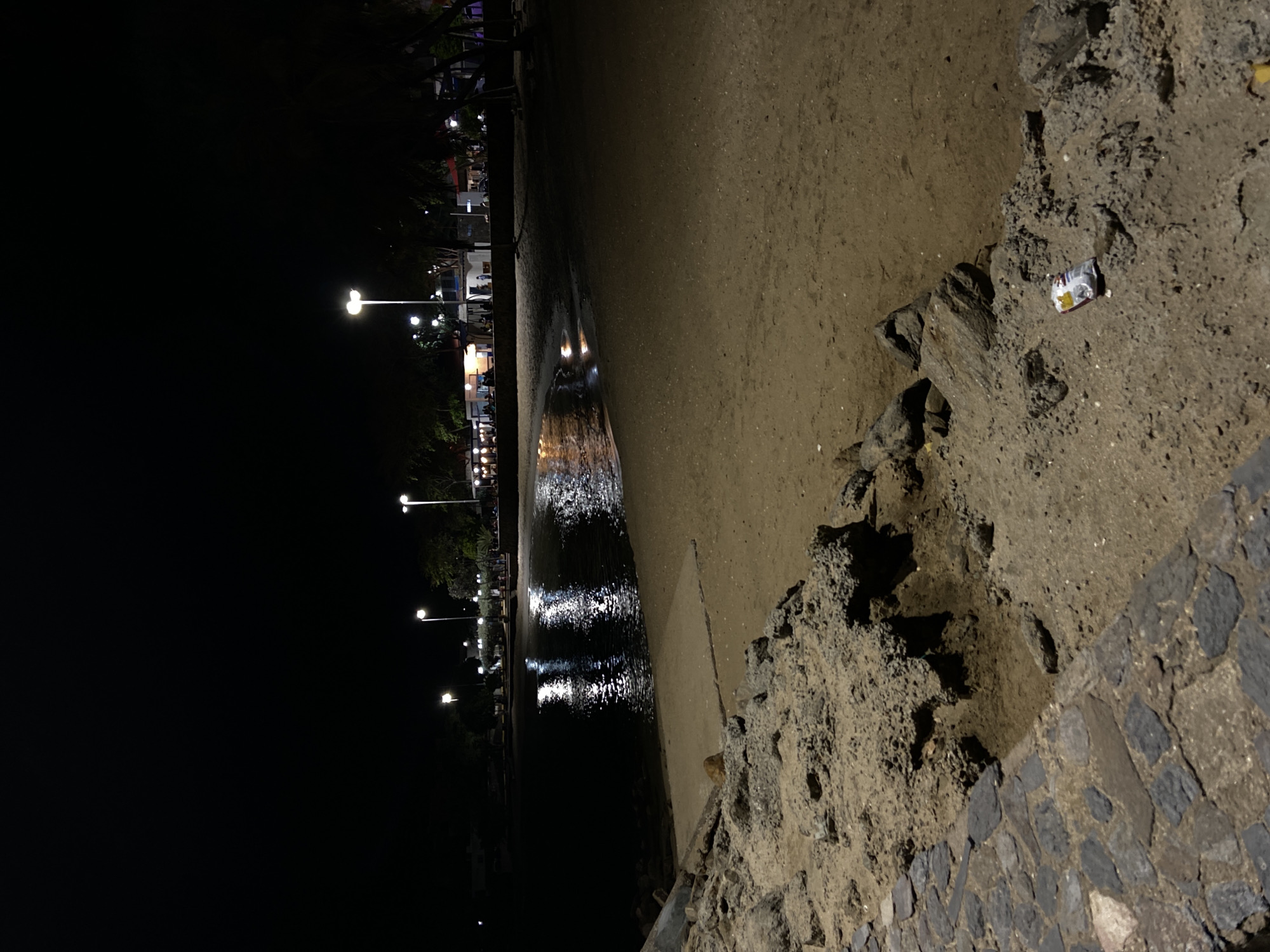 A night scene on the beach in Juan Griego, Venezuela. Streetlights illuminate the shoreline, reflecting in the water. The tide line is visible on the sand. A stone embankment is in the foreground, and litter, possibly a small package, is visible on the ground near the embankment.