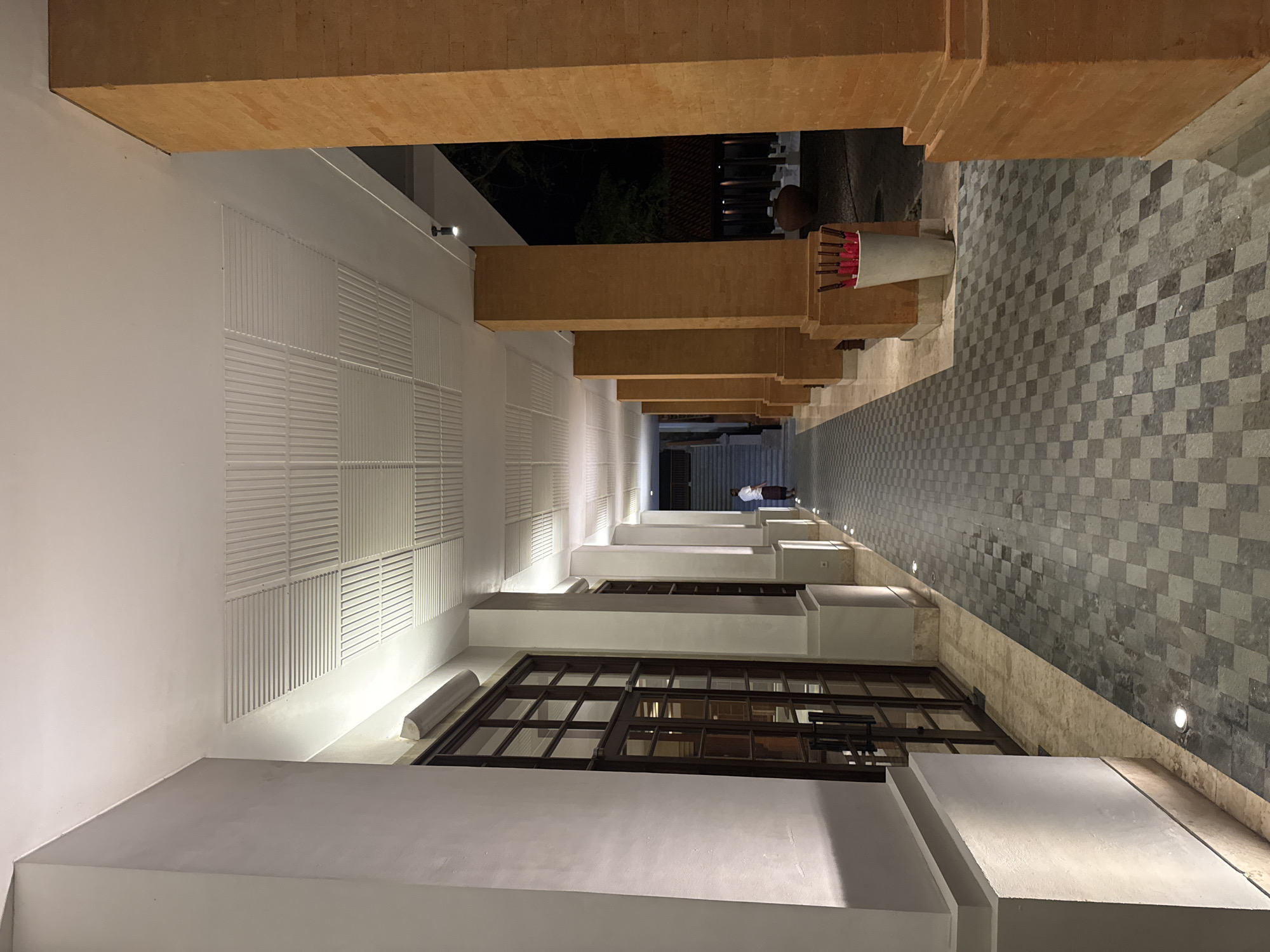 The image shows a view of a corridor with architectural elements in Kuta Selatan, Indonesia. The corridor is defined by white walls on the left and a tiled wall on the right, with wooden beams overhead. The left wall has a series of rectangular, horizontally-slatted designs. Windows are visible, with dark frames and multiple panes. Light fixtures are interspersed along the corridor. A person is visible in the distance. The corridor leads to an open space.