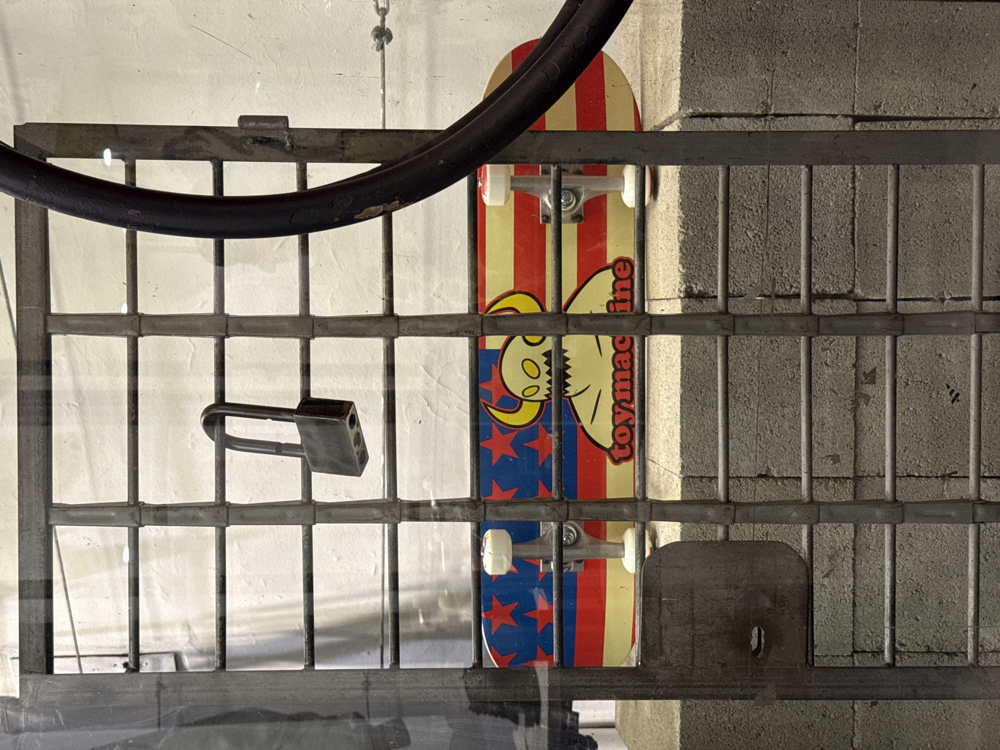 An upside-down close-up captures a skateboard secured behind a metal grate with a padlock. The skateboard is vertical, featuring a red-and-cream striped design with a cartoonish creature and the word "Toymachine." The gate is made of vertical and horizontal bars, and a black metal tube is also visible on the top left. The background behind the skateboard includes a white surface and a concrete wall. A metal plate with a keyhole is secured to the grate in the lower right. The image appears to be taken at an unusual angle, possibly as a reflection. Buenos Aires, Argentina cannot be determined from the image.