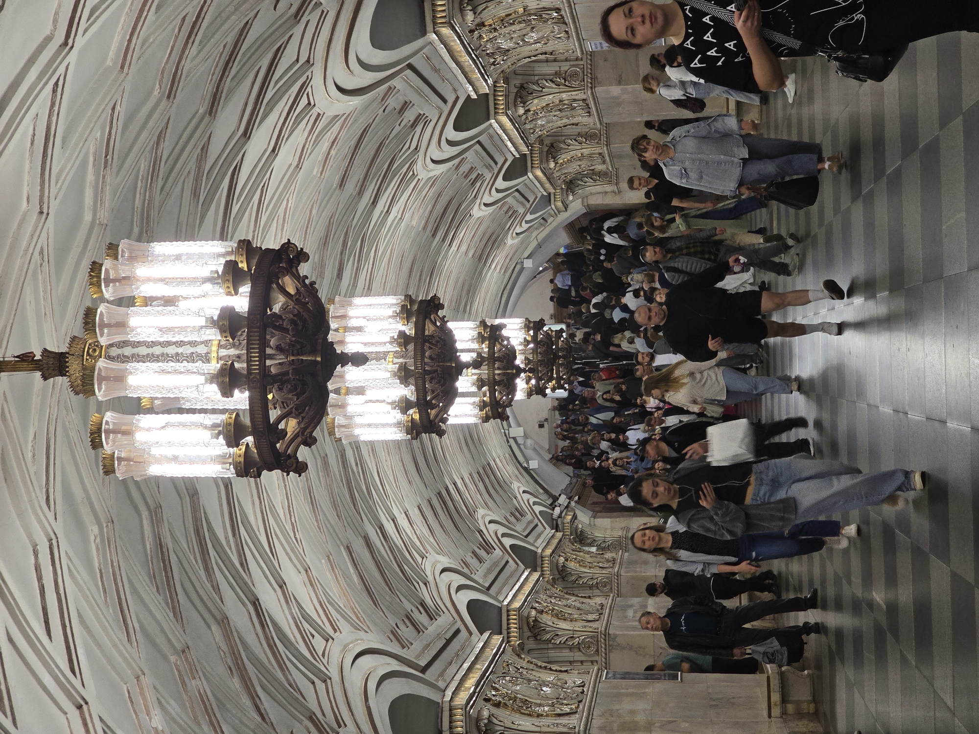 The image captures a crowded scene inside a Moscow metro station in Russia. A line of ornate chandeliers illuminates the vaulted ceiling above a throng of people walking along the platform. The ceiling has intricate plasterwork with repeating arch patterns highlighted with gold trim. The floor is tiled with a grey and white checkerboard pattern. The people in the scene vary in age and attire. Some are looking at their phones, others are talking, and some are simply walking.