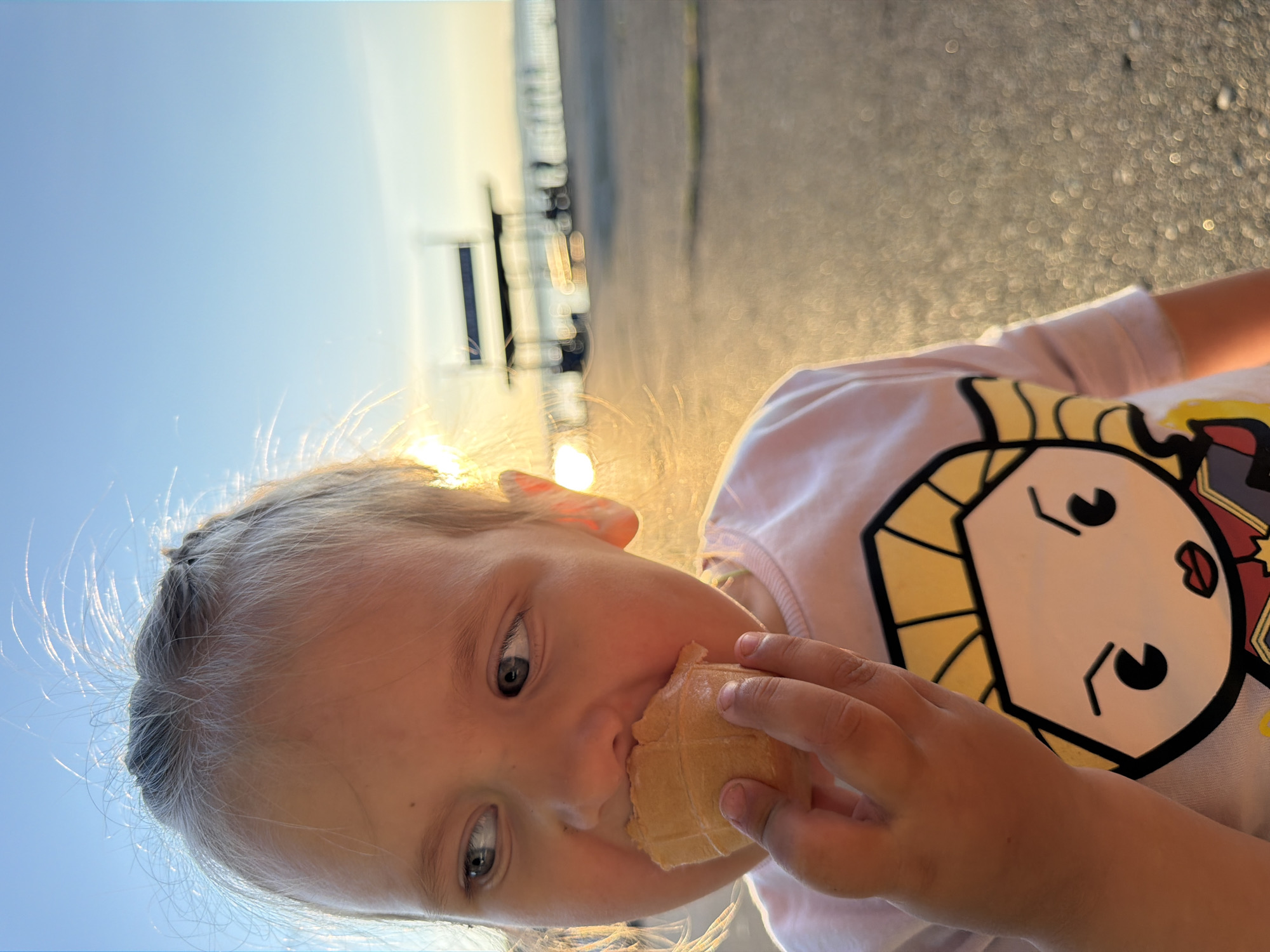 A close-up, low-angle shot shows a young blonde girl eating an ice cream cone in Chaikovsky, Russia. The sun is setting, casting a warm glow on the scene and creating bright highlights. The girl is wearing a pink t-shirt with a cartoon character on it. Her face is turned towards the camera, and she has a serious expression as she eats her ice cream. Her hair is pulled back, with strands of hair catching the sunlight. In the background, a blurred image of a walkway or pier can be seen, with faint lights in the distance. The overall mood is peaceful and contemplative.