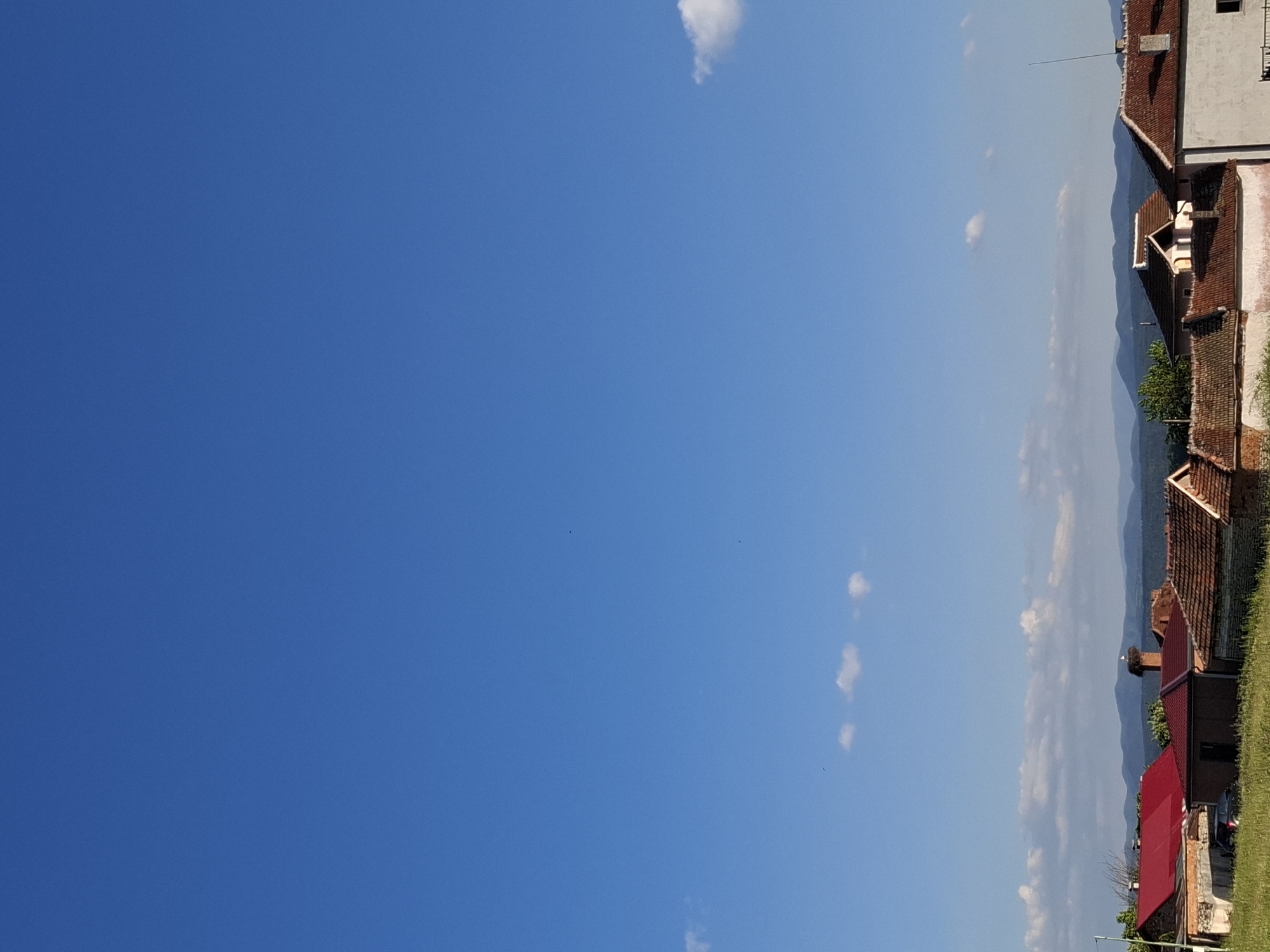 The image presents a scene in Krapets, Bulgaria, featuring a clear blue sky with scattered clouds taking up most of the frame. On the right side, there is a row of buildings with red-tiled roofs, along with some greenery and a glimpse of mountains in the background. The architecture appears to be typical of the region, with houses constructed from brick and white painted walls.