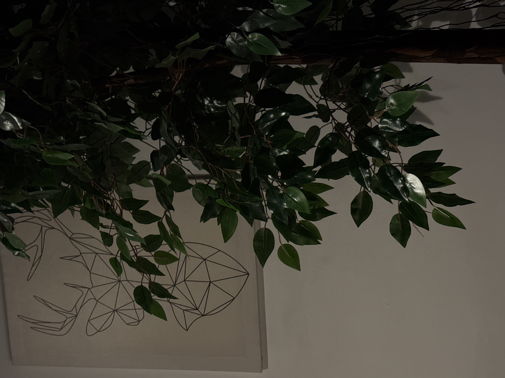The image shows a canvas with a geometric deer outline on a plain wall, partially obscured by the branches and leaves of a large artificial plant. The plant has dark green, glossy leaves and a dark brown trunk. The lighting is dim, casting shadows on the wall. The scene appears to be indoors, likely in a residential or commercial setting. There are no people or animals visible in the image. The location in the city of Chaikovsky, Russia, is not discernible from the visual information provided.
