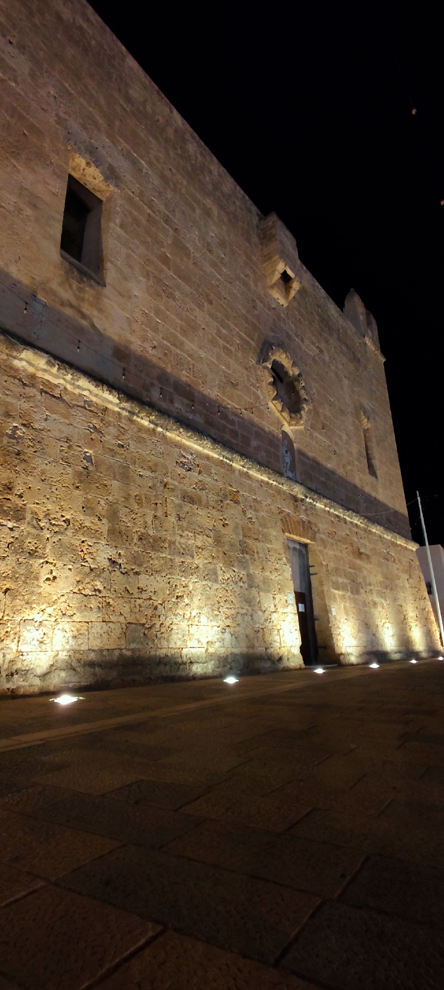 The image shows a stone building, likely a historical structure, at night in San Vito Lo Capo, Italy. The building is illuminated by several ground-level spotlights, casting strong upward shadows on the textured stone walls. The architecture appears to be old, with large, roughly cut stones forming the facade.
Key architectural features visible include:
* A narrow, recessed window on the upper left portion of the building.
* A prominent circular decorative element or window opening with intricate stonework, located on the upper central part of the facade.
* A diamond-shaped opening above the circular feature.
* A small, sculptural figure positioned near the base of the wall, below the circular opening.
* An arched doorway, partially obscured by a door, on the right side of the visible facade.
* A string of small, globe-shaped lights is suspended along a horizontal band running across the middle of the building's facade.
The ground in front of the building is paved with large, rectangular stone slabs, also illuminated by the ground lights, creating distinct patterns of light and shadow. The sky is dark, indicating it is nighttime. The overall impression is one of a well-preserved historical site illuminated for nighttime viewing.