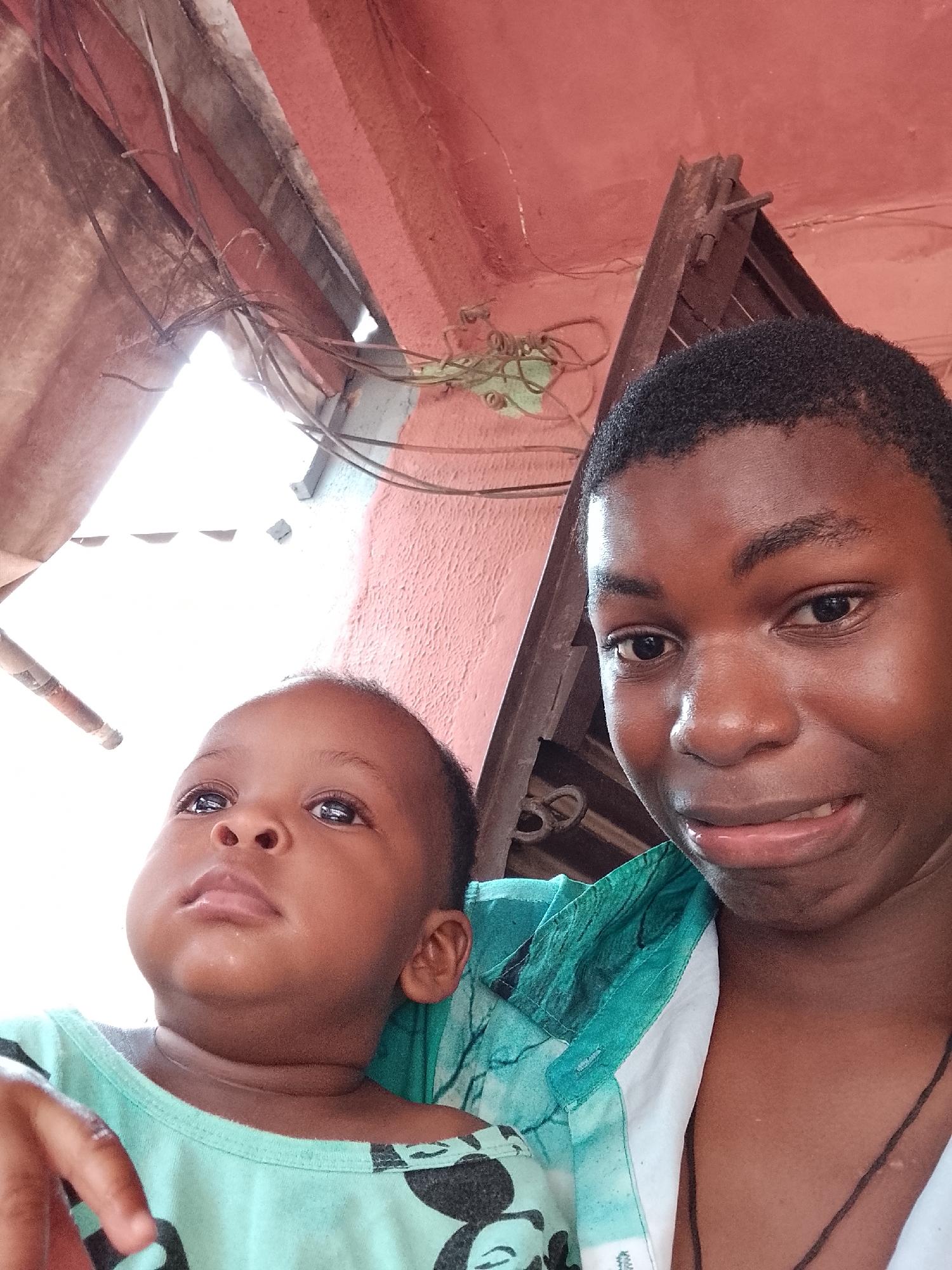 A candid portrait features two individuals: an adult or older teenager on the right, holding a baby or toddler on the left. The adult, with dark skin and short, dark, curly hair, looks directly at the camera with a subtle smirk. They are wearing a shirt patterned in white and various shades of green.
The baby, also with dark skin and short, dark hair, looks upwards and slightly to the left, with a contemplative expression. They are dressed in a light green shirt adorned with an indistinct dark pattern, possibly a cartoon character.
The setting appears to be an indoor or semi-sheltered outdoor space, characterized by a reddish-orange ceiling or upper wall. Tangled dark wires or roots are visible in the upper left, suggesting electrical wiring or plant growth in an older structure. Behind the adult's head, part of a rustic wooden or metal frame is discernible. A bright, overexposed light source, likely a window or open area, dominates the upper left background. The overall lighting suggests daytime. No visible text is present in the image.