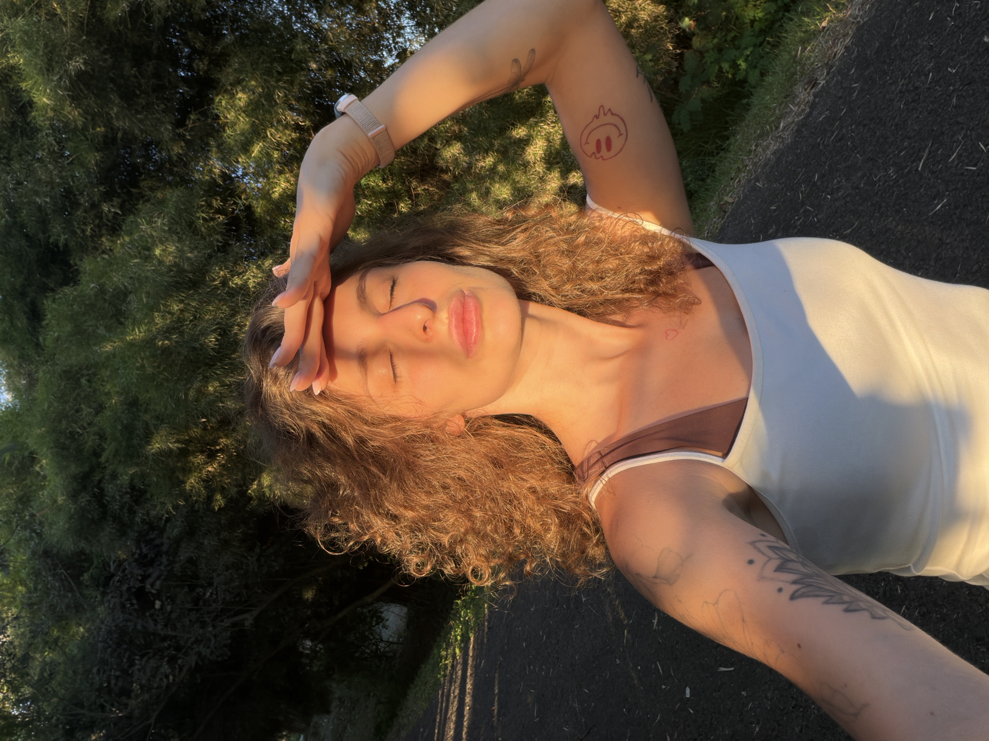 A woman with curly brown hair and closed eyes stands outside, shielding her face from the sun with her left hand. She wears a white tank top and a brown sports bra underneath. The sun shines on her face, shoulders, and arms. She has tattoos on both arms, one with a cartoon-like face and another with a more complex design. She also has a watch on her left wrist. A small heart tattoo is visible on her neck. The background consists of trees and a paved surface. The location appears to be a park or green area. The image does not specify the location in the city of Kediri, Indonesia.