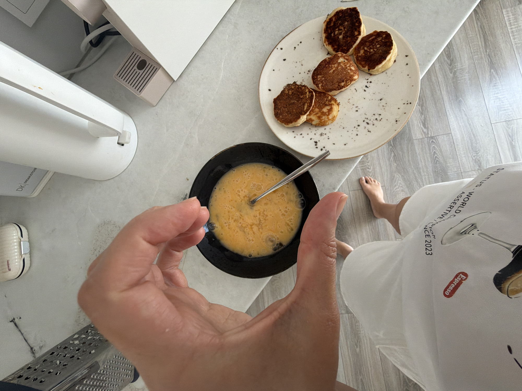 In Malinovka, Russia, a person's hand is positioned over a black bowl containing whisked eggs. To the right, a white plate holds several small, golden-brown pancakes. The background shows a countertop with kitchen appliances, including a grater, a white kettle, and a pink decorative object. The floor is tiled in a light gray wood-grain pattern. The person is wearing a white garment with text and an image of a wine glass. The scene appears to be a domestic setting, possibly a kitchen.