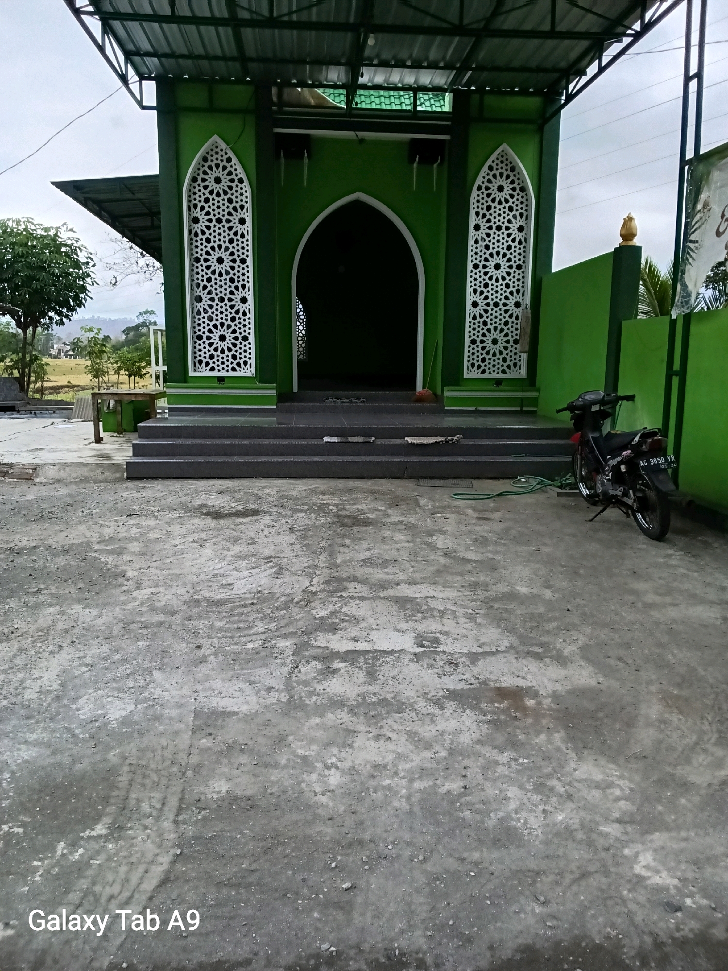 The image shows a green building with ornate white latticework windows. The building appears to be a mosque or a similar religious structure, with steps leading up to an arched entrance. In front of the building, there is a paved area with a motorcycle parked on the right side. The environment suggests an outdoor setting, possibly in a rural or semi-urban area, with trees and hills visible in the background. The sky is overcast, indicating a cloudy day. The text "Galaxy Tab A9" is present in the bottom left corner of the image. The license plate on the motorcycle reads "AG 3850 YR". Based on the location of Trenggalek, Indonesia, this scene likely depicts a local place of worship or community building.