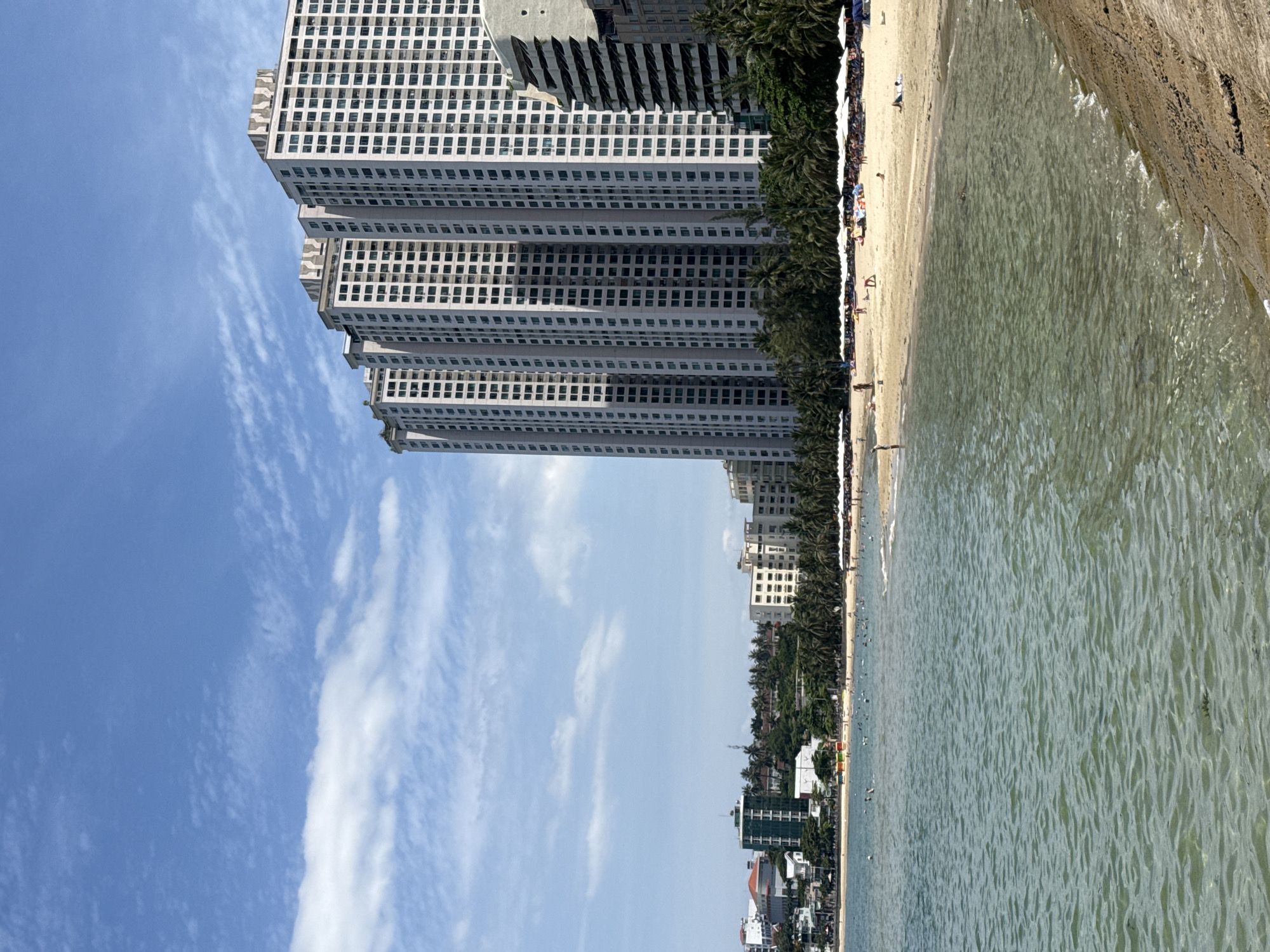 The image depicts a bustling beach scene in the city of Bac Nha Trang Ward, Vietnam, under a bright, partly cloudy sky. A series of tall, modern apartment buildings dominate the background, their white facades and numerous windows contrasting with the blue sky. In front of the buildings, a sandy beach stretches along the coastline, lined with rows of white umbrellas and beach chairs. Numerous people are visible on the beach, some sunbathing, others walking or engaging in leisure activities. The ocean in the foreground is a clear, turquoise color, with small waves lapping at the shore and a concrete embankment. Several individuals are in the water, swimming or wading. The overall impression is one of a vibrant, popular tourist destination.