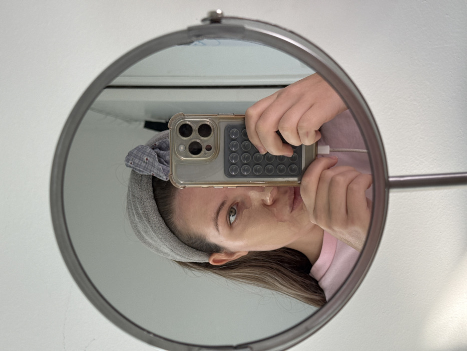 A female subject is seen taking a mirror selfie in an indoor setting within Vozdovac Urban Municipality, Serbia. Her reflection occupies the majority of a round, silver-framed vanity mirror. The subject has long, brown hair pulled back by a grey terrycloth headband, with a small, plaid-patterned fabric accessory positioned above her right ear. Patches of a beige substance, consistent with a facial mask or incomplete makeup, are visible on her face. She wears a light pink crew-neck shirt. Both hands hold a clear-cased smartphone, identified as an iPhone, which has a grey accessory featuring multiple circular suction cups on its back. A white charging cable is connected to the phone. The environment consists of a plain white wall visible behind the mirror.