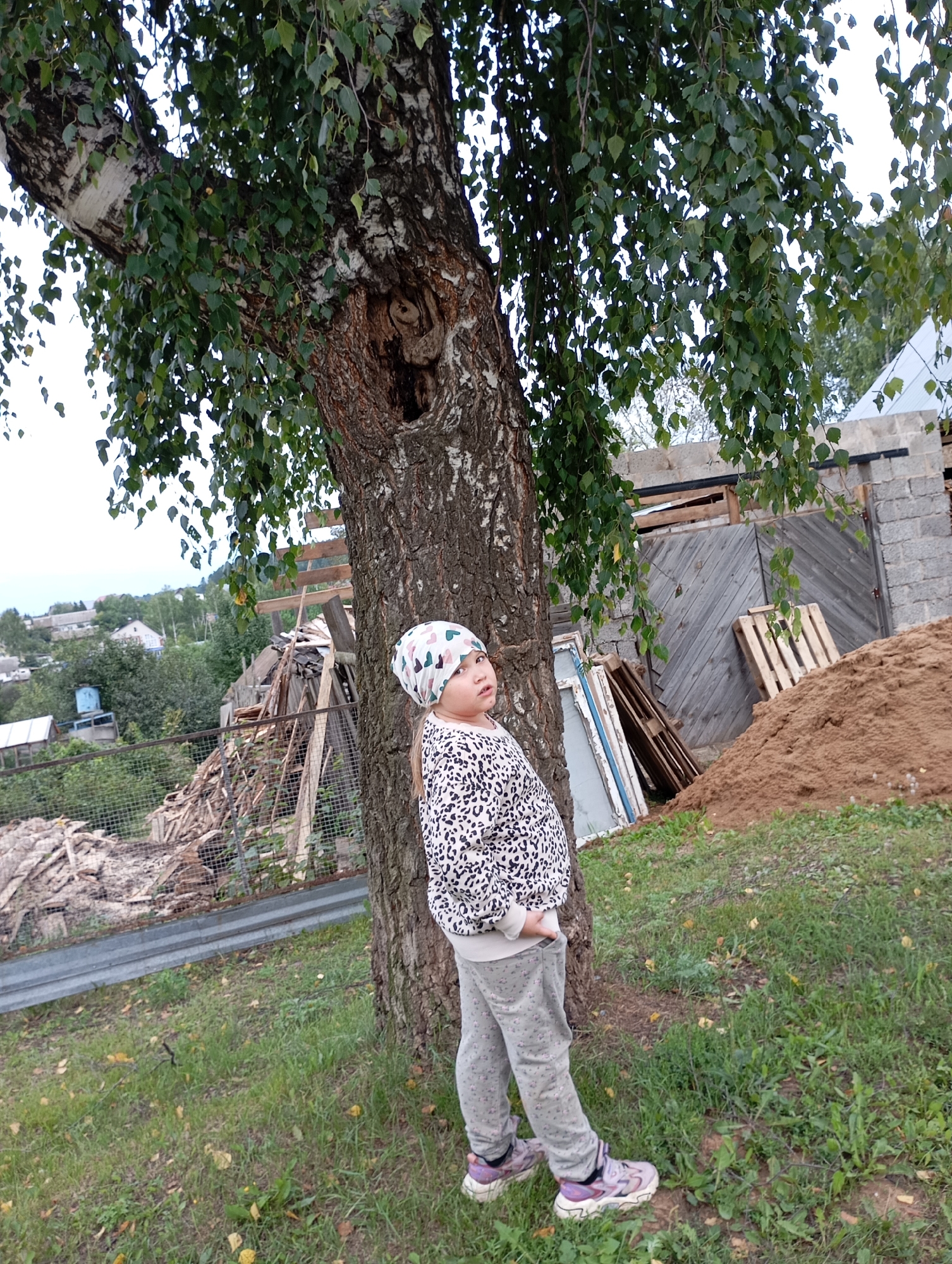 A young girl with blonde hair and a patterned headscarf stands next to a large tree. She is wearing a leopard-print sweatshirt and grey sweatpants with a floral pattern, and purple sneakers. The girl is looking up and to her left. The tree has a large hole in its trunk. Behind the girl and the tree, there is a pile of sand, wooden pallets, a wooden fence, and a building under construction. In the distance, there are houses and trees, suggesting a rural or semi-rural setting. The sky is overcast. The image appears to be taken in the city of Fokino, Russia.