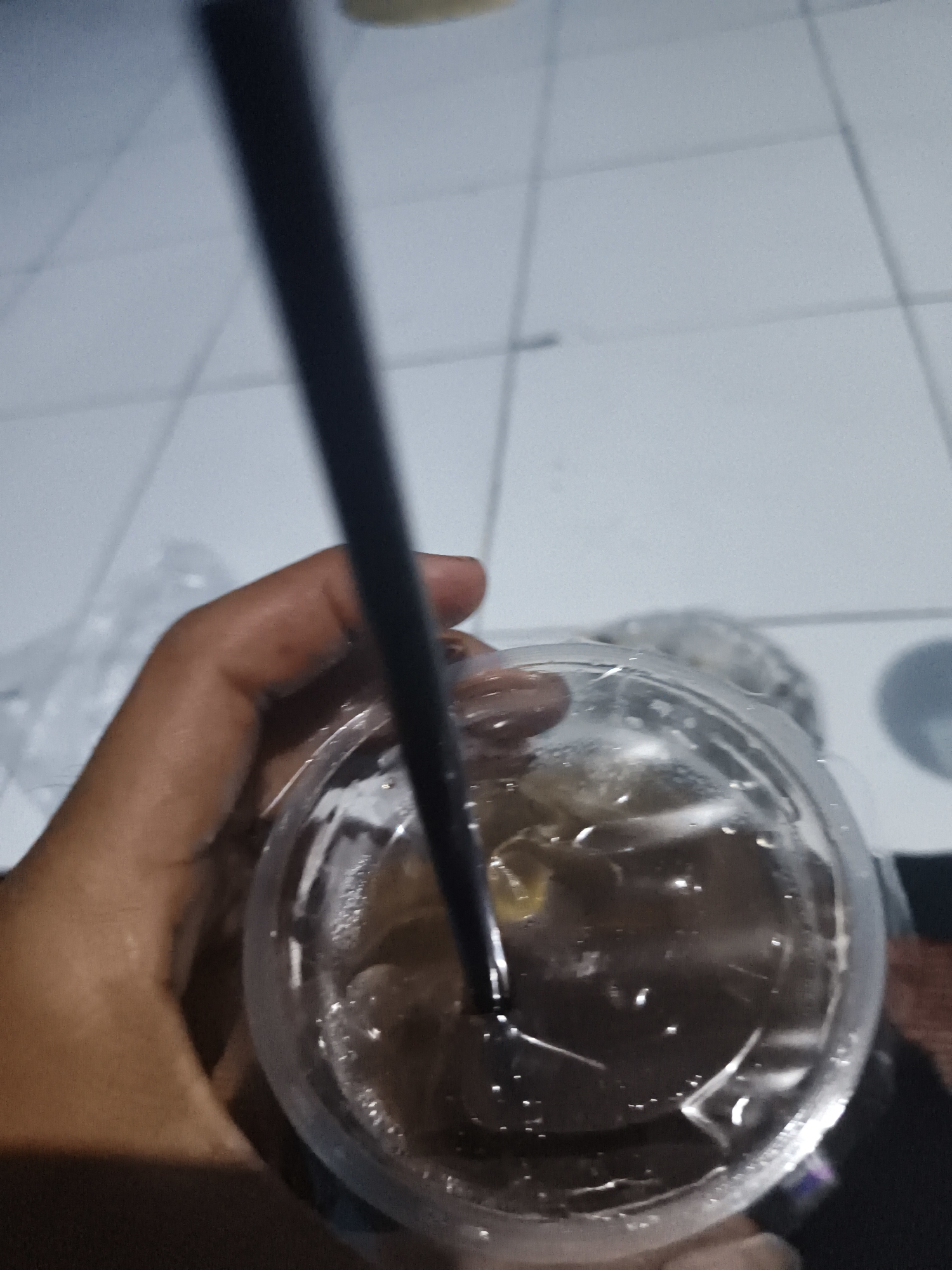A person's hand is holding a clear plastic cup containing a dark liquid with ice cubes and a black straw. The cup is partially covered with a plastic lid. The background shows a tiled floor with faint grid lines. The image appears to have been taken indoors. The exact location within Pangkalpinang, Indonesia, cannot be determined from the visual information provided.