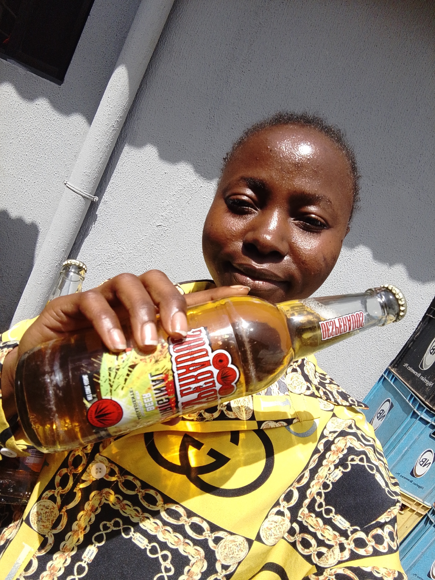 The media file shows a close-up selfie of a woman holding a bottle of beer. She is looking towards the camera with a slight smile. The woman is wearing a yellow shirt with a black and gold chain pattern. The bottle she is holding has a label that says "Pilsner Lager Beer" and appears to be a Nigerian beer. In the background, there are stacked crates, possibly of beer bottles, and a grey textured wall with a pipe. The lighting suggests it is daytime, likely outdoors. The woman's face is shiny, indicating sweat, which could suggest warm weather. There are shadows cast on the wall, indicating the sun is out. The overall scene appears to be casual, possibly a social gathering or a moment of relaxation. No specific time of day is discernible beyond it being daylight. The location context is Jos, Nigeria.