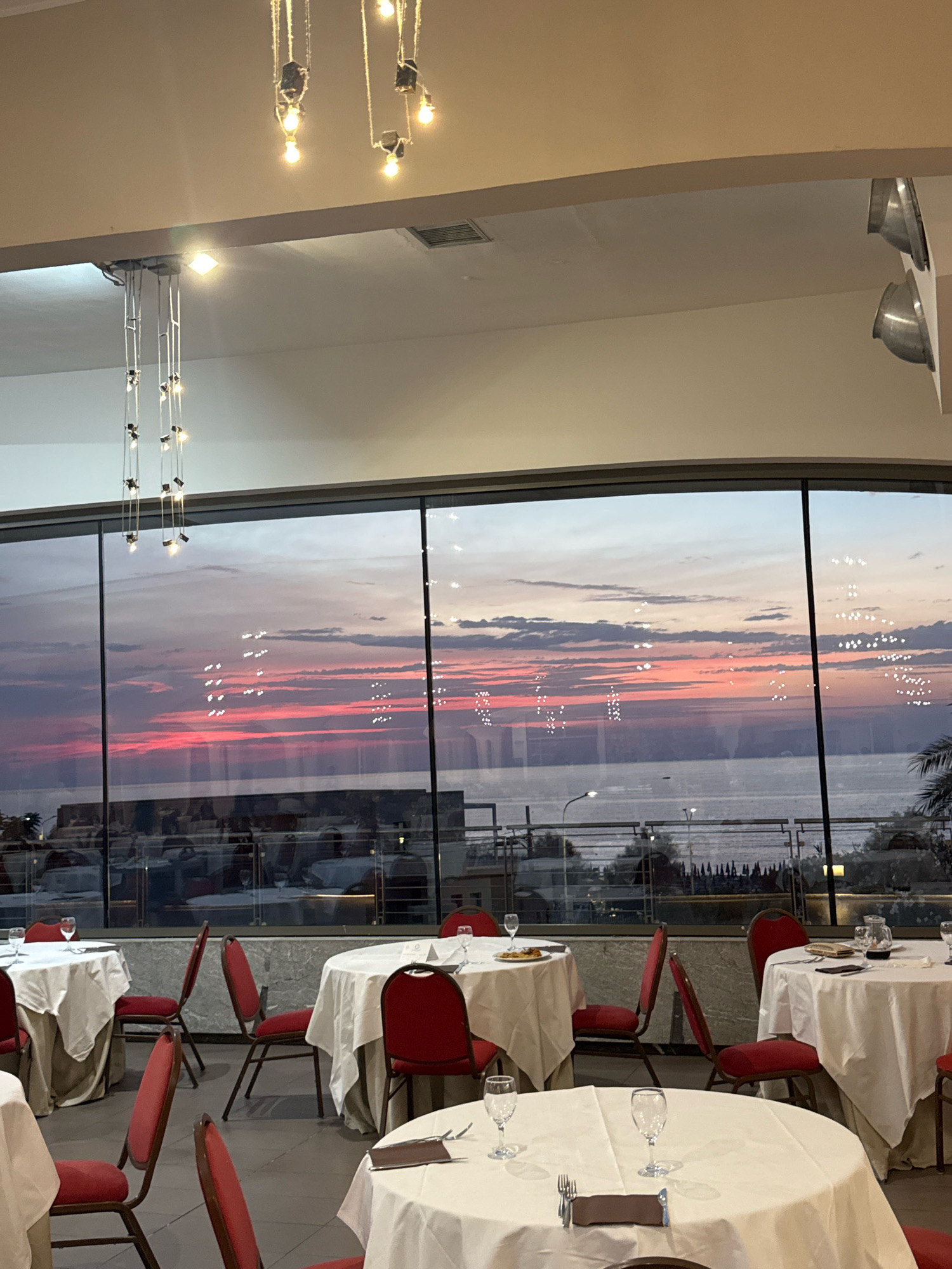 The image depicts the interior of a restaurant, likely in Capaci, Italy, with a large window offering a view of the sea and a vibrant sunset. The restaurant is set up for dining with round tables covered in white tablecloths and surrounded by red chairs. Several tables are visible, some with place settings including glasses and napkins. In the foreground, one table has silverware and a napkin. The window shows a wide expanse of water with a colorful sky filled with clouds painted in hues of pink, orange, and purple. Reflections of interior lights are visible on the glass. Outside the window, a railing and some trees can be seen along the waterfront. The ceiling features modern pendant light fixtures. The overall scene suggests an upscale dining establishment with a picturesque view.