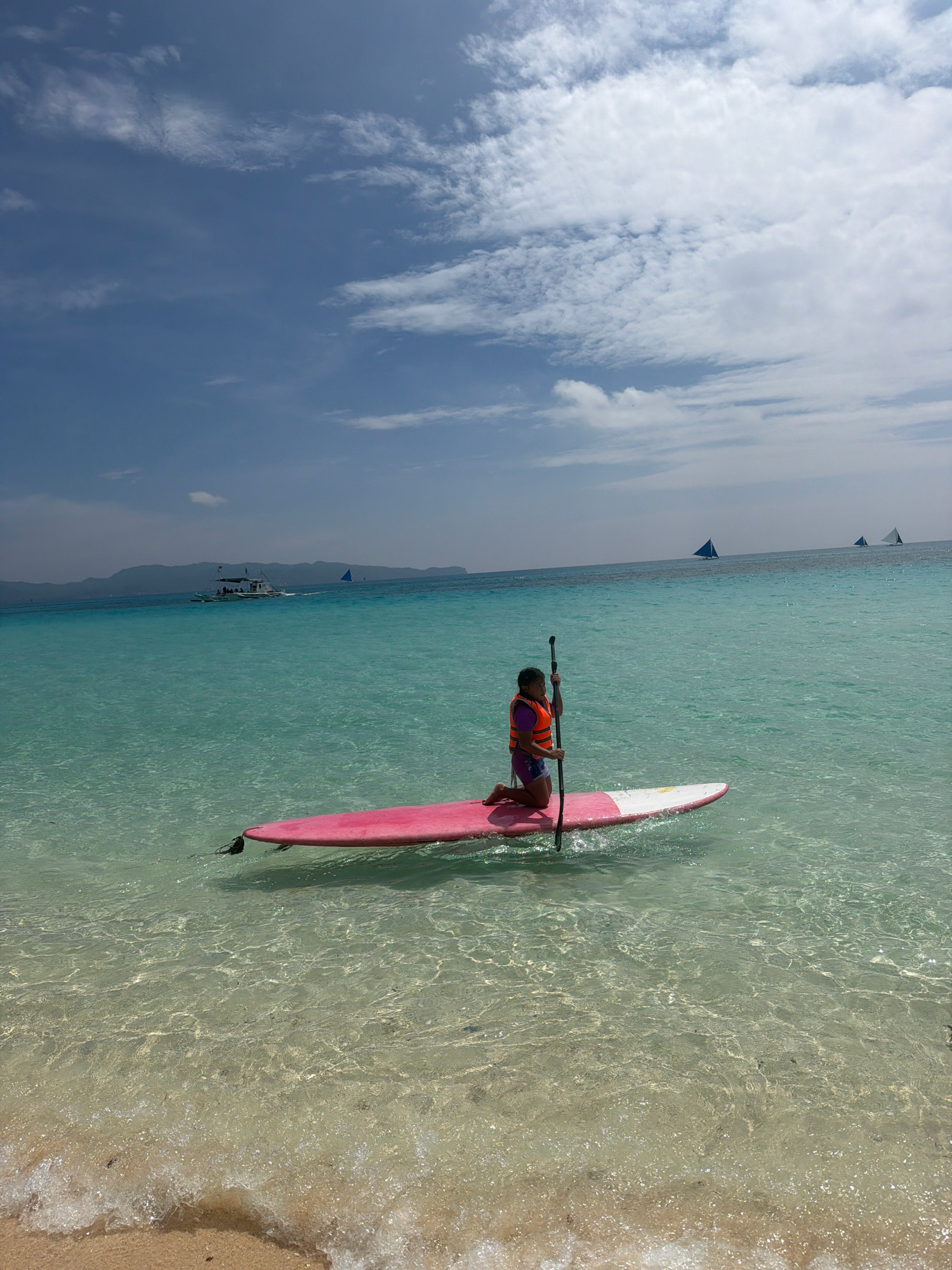 A young girl in a life vest kneels on a pink and white paddleboard in shallow, clear turquoise water. She holds a paddle upright. The water is so transparent that the sandy bottom is clearly visible, with rippling sunlight creating patterns on the seabed. In the distance, a large white boat with people on board is visible, along with a few sailboats with triangular sails. The sky above is a light blue with scattered white clouds. The scene appears to be a tropical beach, likely in the city of Malay, Philippines, known for its beautiful waters.