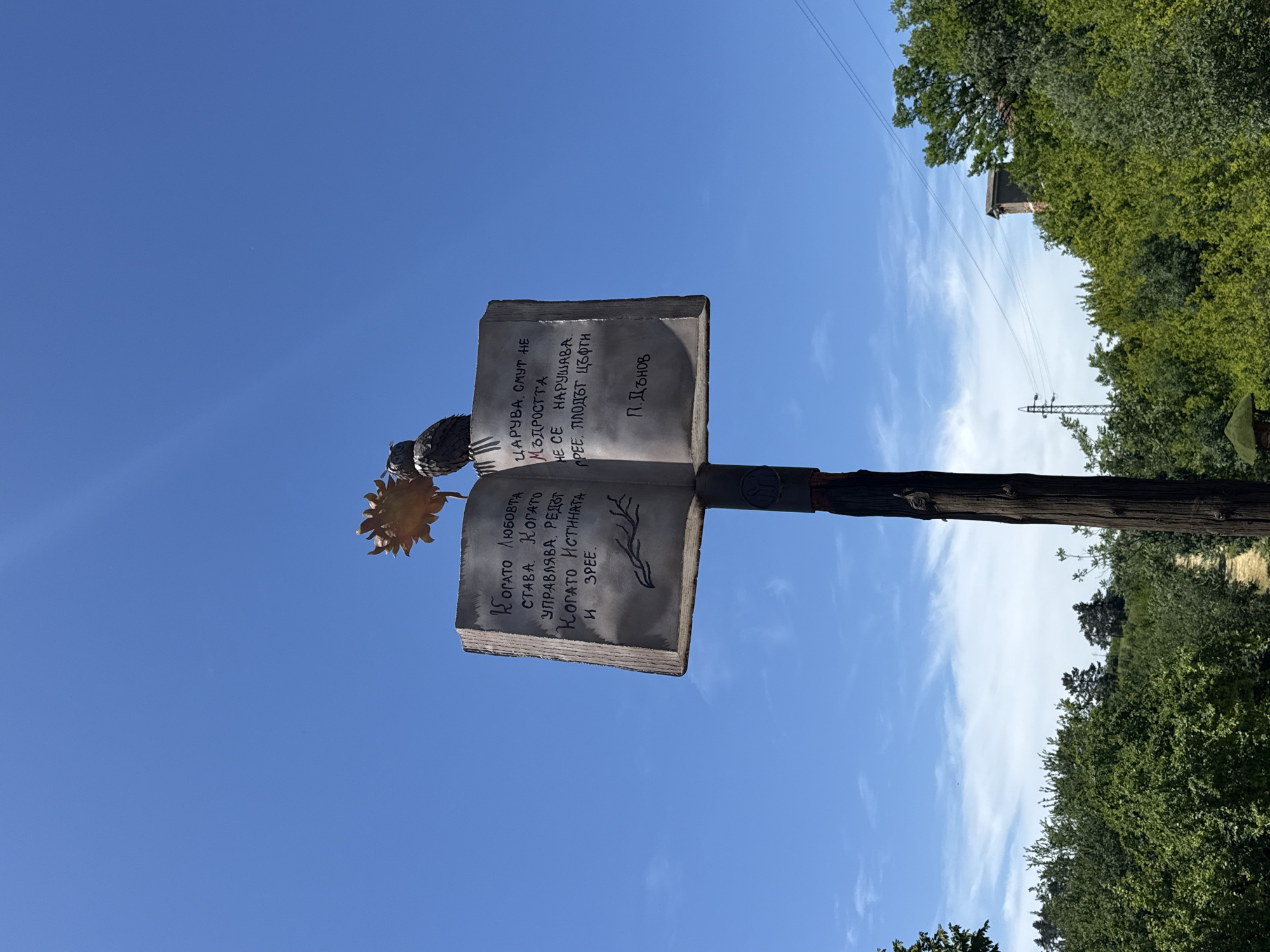 The image shows a metal book sculpture atop a wooden post. The book is open, and Bulgarian text is inscribed on both pages. An art piece resembling a sunflower is attached to the side. The location is outdoors, under a bright blue sky with scattered clouds. In the background, greenery and trees suggest a park or natural environment. Based on the text in the book, this is the Park Iskar Panega in Lukovit, Bulgaria.