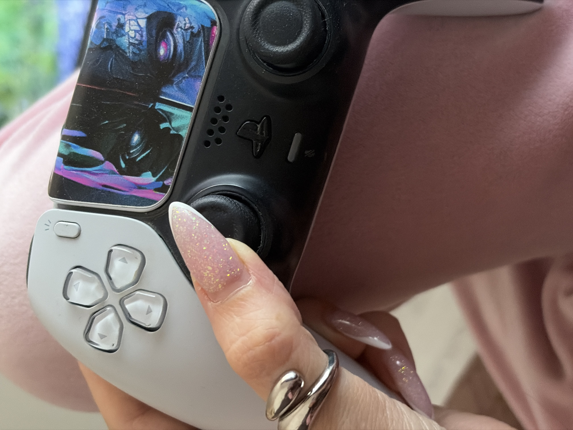 The image shows a close-up of a person's hand holding a PlayStation DualSense controller, suggesting engagement in video gaming. The controller, primarily white and black, features a custom, artistic decal on its touchpad. This decal depicts an abstract, dark scene with dominant blue, purple, and black colors, reminiscent of a fantastical or science fiction landscape.
The hand has long, manicured nails, shaped in a coffin or almond style, painted with a light pink, glittery polish. Some nails also display white French tips. A distinctive silver-toned, wrap-around ring is worn on the finger operating the right analog stick.
Key controller features visible include the white directional pad (D-pad) with clear buttons indicating arrows, a partially visible PlayStation logo on the home button, a mute button, and a series of small microphone speaker holes. The background is softly blurred, revealing hints of a light pink or peach-colored fabric, likely clothing or a blanket, and indistinct green and blue blurs that could be from a screen or an outdoor view. The setting is indoors, with no discernible time of day or specific weather conditions. While the provided context is Saint Petersburg, Russia, the image itself offers no specific geographical cues for this location. No other explicit text is visible.