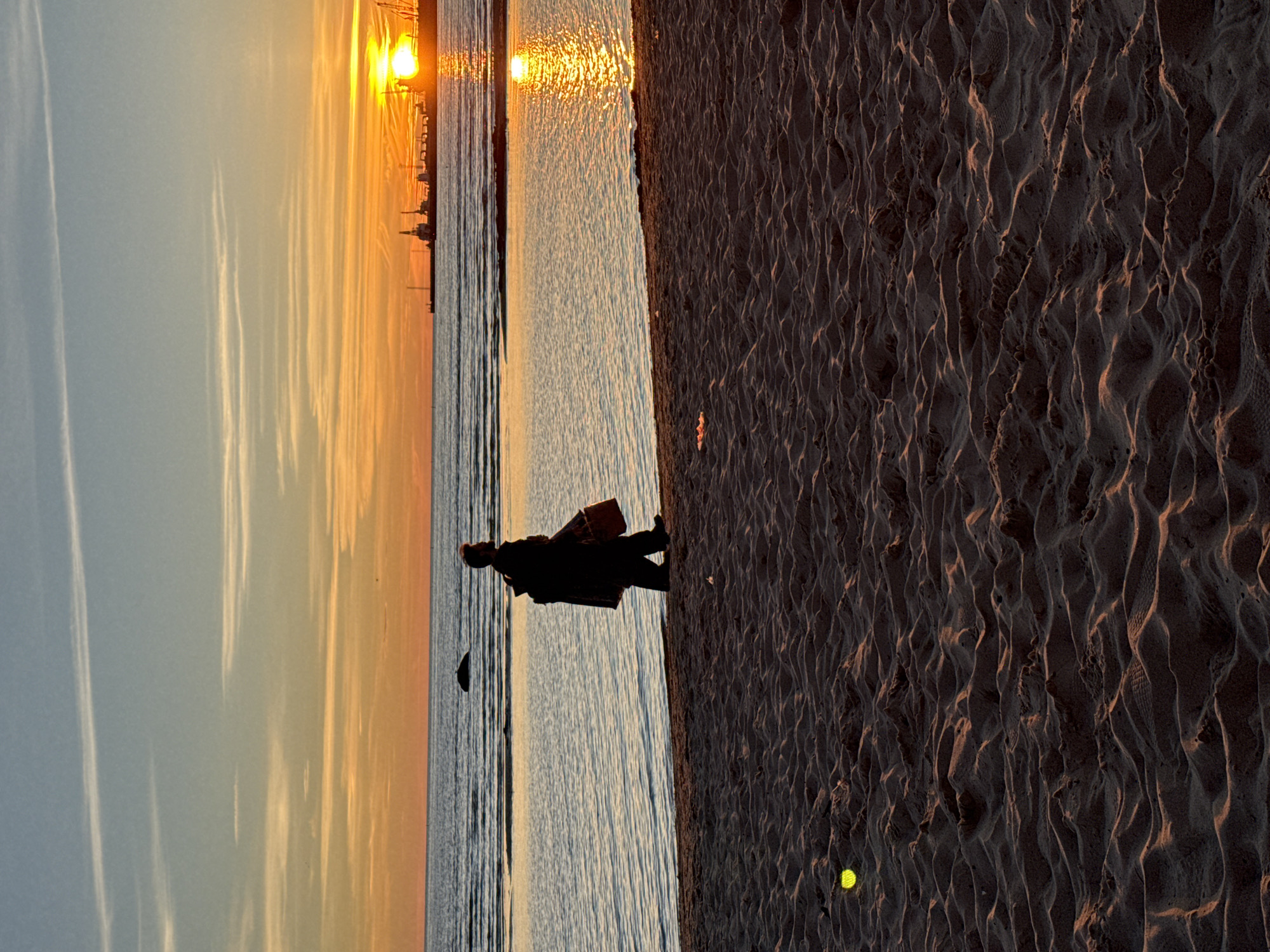 This is a photograph taken at sunset on a beach in Tallinn, Estonia. The sun is setting over the Baltic Sea, casting a warm, orange glow across the sky and water. In the foreground, a person is walking along the sandy shore. The person is silhouetted against the bright sky, and appears to be carrying a bag. In the background, the silhouette of a port with cranes and ships is visible on the horizon. The sand in the foreground is rippled, suggesting it is a beach. There is a small, bright yellow object on the sand in the lower left quadrant of the image, possibly a tennis ball or a similar object. The overall mood of the image is serene and peaceful.