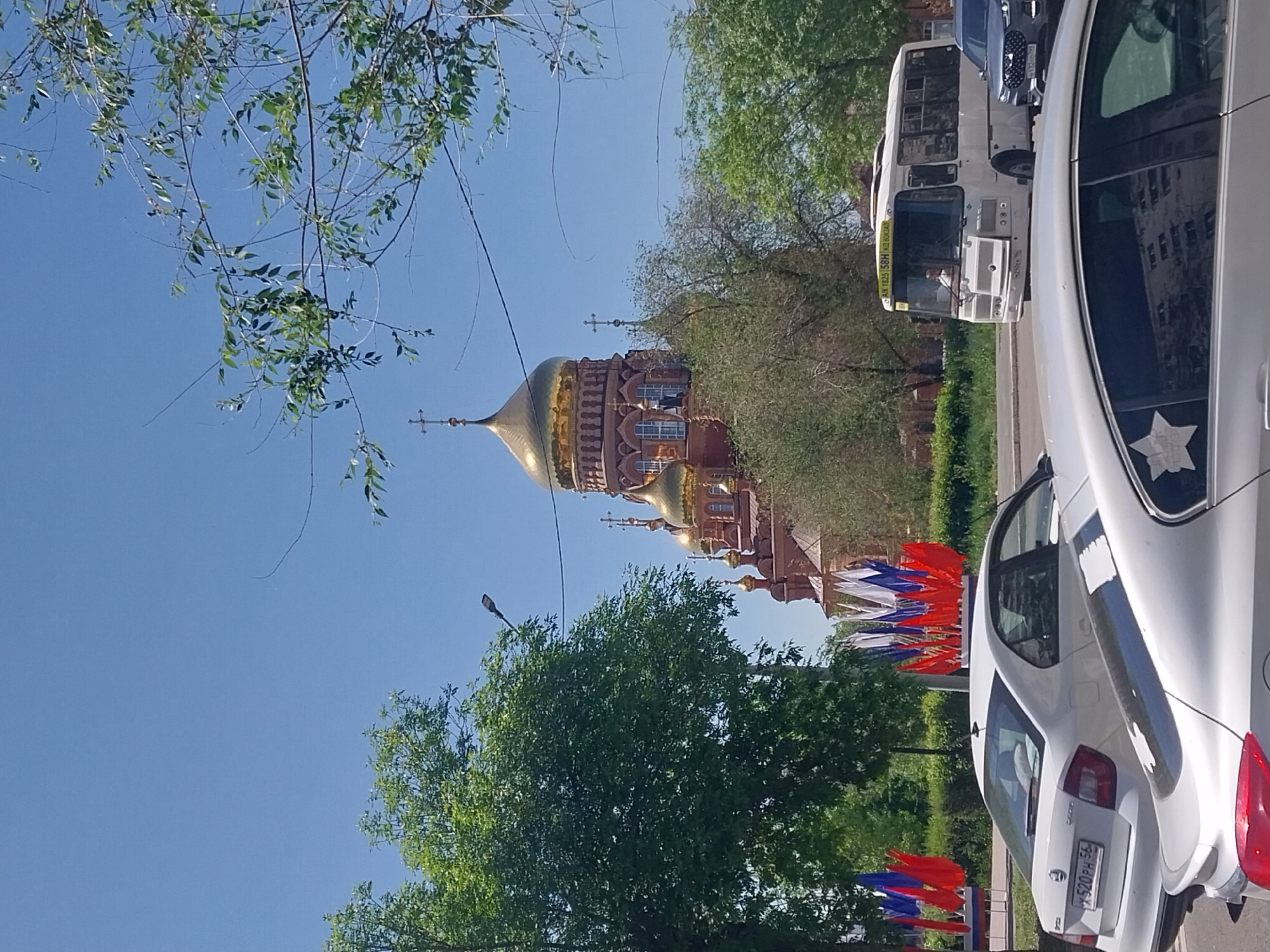 The image depicts a view of Orenburg, Russia. The focal point is a large, ornate Orthodox church with multiple golden domes. The church is constructed of red brick. In the foreground, a white car is parked on the right side of the frame. A white bus is behind the car. The street is lined with trees and Russian flags. The sky is clear and blue.