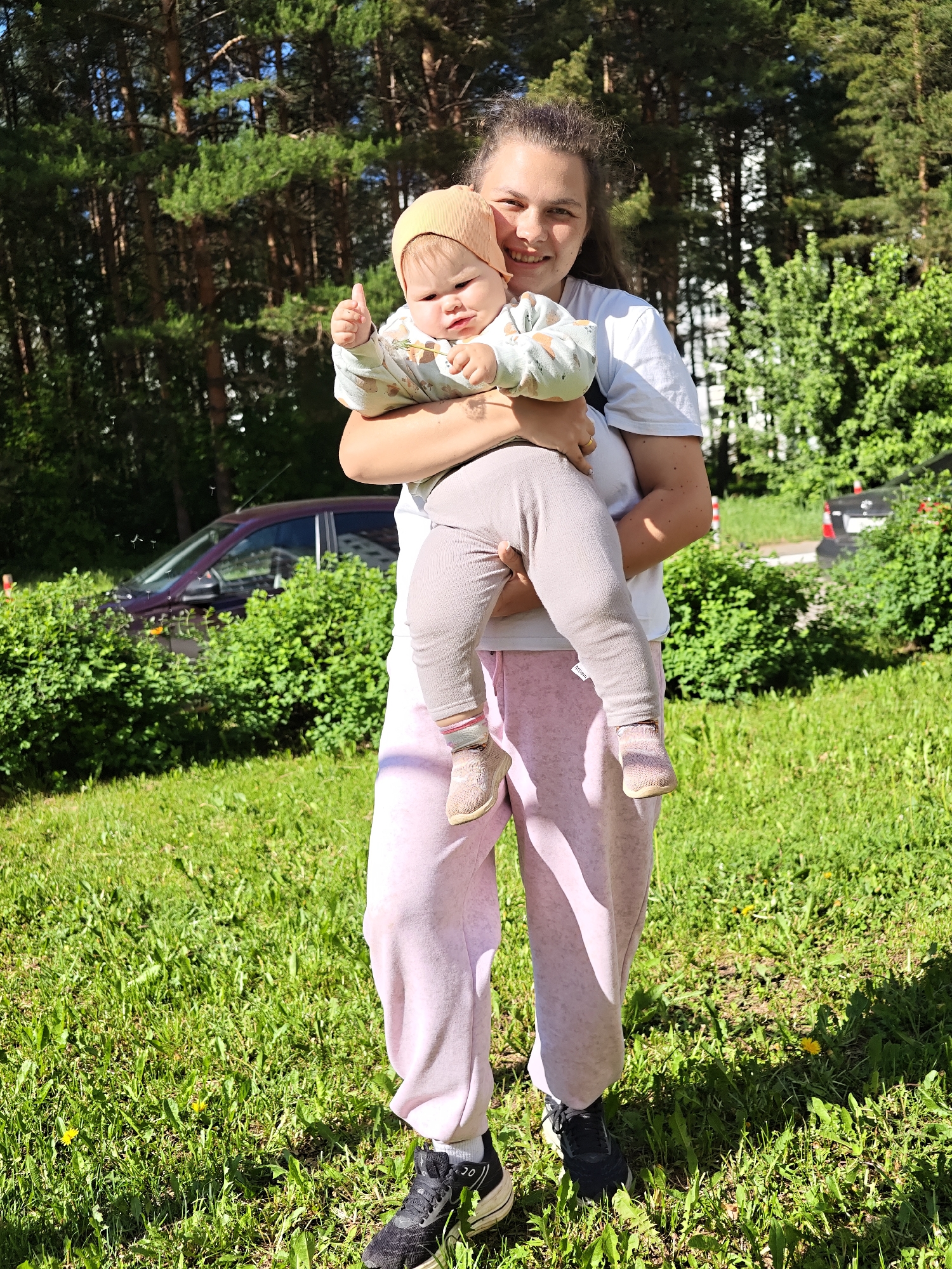 The image shows a young woman holding a baby in a grassy outdoor setting. The woman has long brown hair pulled back, wearing a white t-shirt and light pink sweatpants with dark sneakers. She is smiling. The baby is wearing a light-colored patterned top, beige pants, a beige hat, and light pink shoes. The baby's left hand is raised giving a thumbs-up. In the background, there are trees, bushes, and a dark red car is partially visible behind the bushes. The location is in Chaikovsky, Russia. The scene is sunlit and appears to be taken during the day.