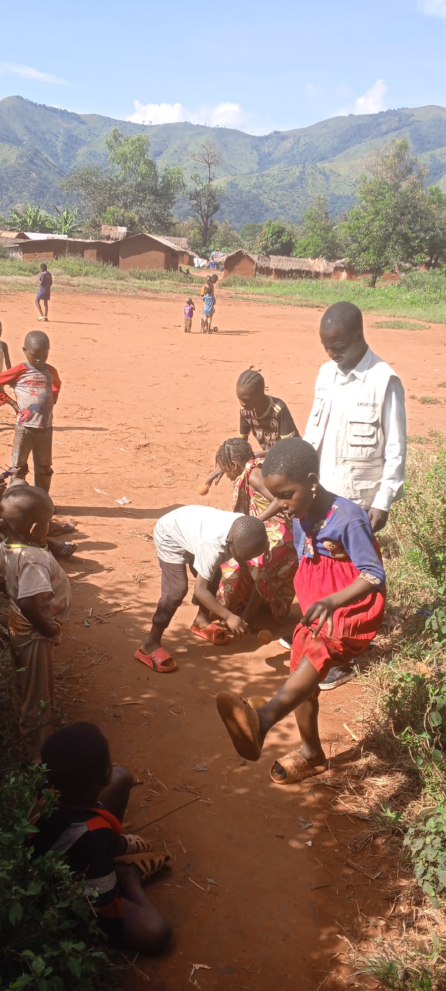 In Ngalula, Democratic Republic of the Congo, a group of children and an adult man are on a dirt path in a rural area. Several children gather around, some bending over and appearing to interact with objects on the ground. One child wears a blue and red dress and raises a leg. An adult man in a light-colored collared shirt and vest stands nearby, seemingly observing the children. In the background, there are simple, mud-brick structures with thatched roofs, and further off, rolling hills and mountains are visible under a bright, clear sky. The ground is reddish-brown dirt, and there is some sparse vegetation along the edges of the path. Other children are dispersed in the background.