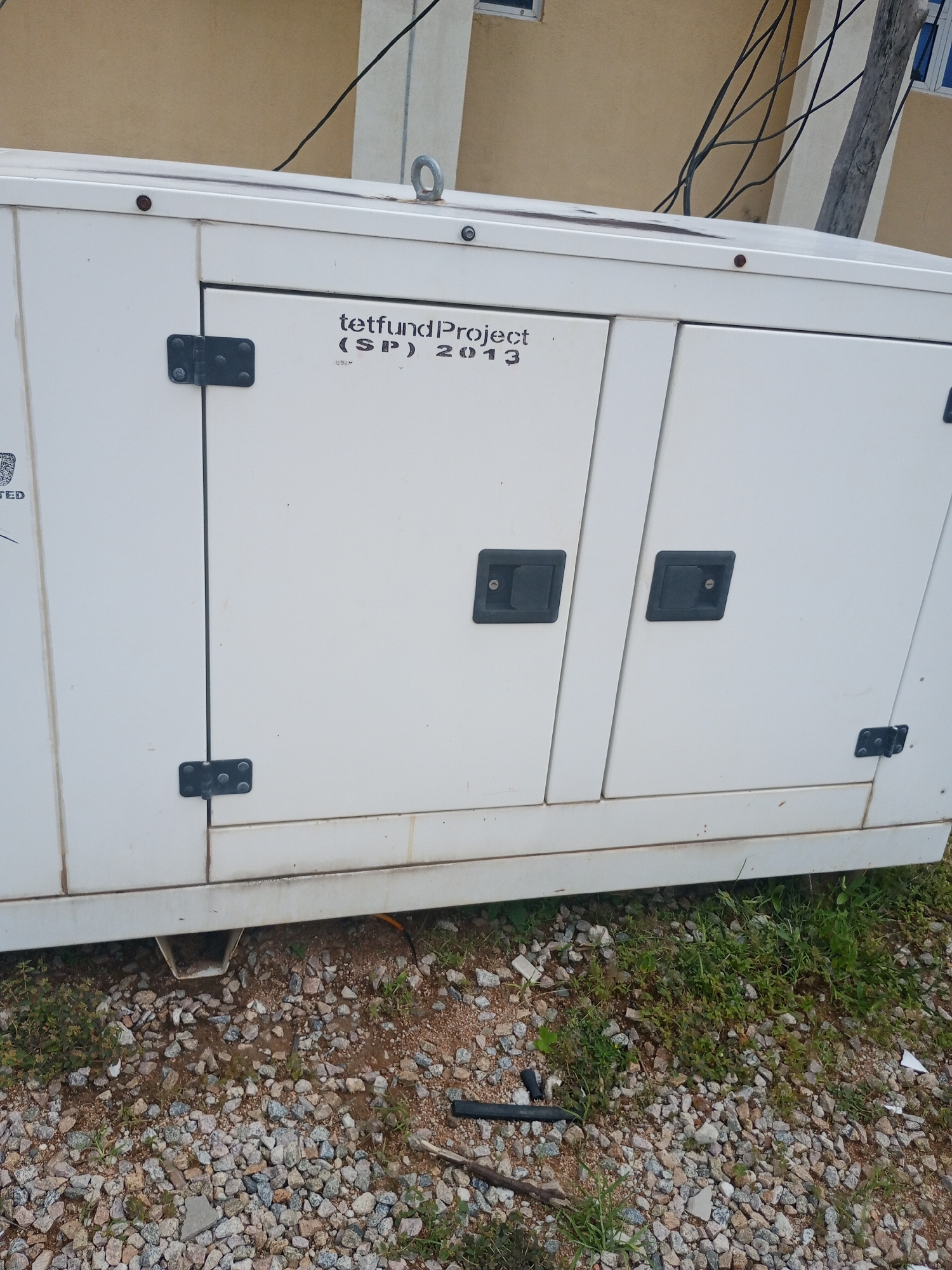 The image shows a white generator located outdoors on a gravel and dirt surface with some grass and small plants growing. The generator is a large, rectangular unit with two doors on the front. The left door has a black handle and hinges, and a small sticker with text is visible on the far left. The right door also has a black handle and hinges. Above the doors, printed in black, is "tetfund Project (SP) 2013". Overhead, black electrical wires are strung across the scene, and a building with a light brown facade and white trim is visible in the background. A wooden pole is also visible to the right of the building. The ground beneath the generator appears to be covered in small, greyish-brown gravel and some reddish-brown dirt, with a patch of green grass to the left and right. There is a small, white plastic chute protruding from the bottom of the generator, angled towards the ground. The overall scene suggests an outdoor placement for the generator, possibly associated with the building in the background. The image appears to have been taken in Jalingo, Nigeria.