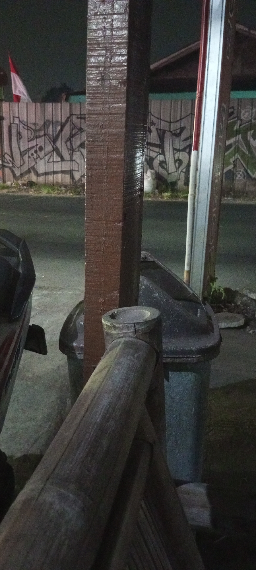 The image depicts a street scene at night, with a focus on a wooden post in the foreground. Behind the post, a trash bin is visible, as is a portion of a motorcycle on the left. The background features a corrugated metal fence covered in graffiti, and an Indonesian flag is flying on the left side. A striped pole, possibly a street light or signpost, stands behind the wooden post on the right. The ground appears to be asphalt, suggesting a road or parking area. The overall impression is one of an urban environment at night, possibly in Setu, Indonesia, given the presence of the Indonesian flag and the general context of the scene.