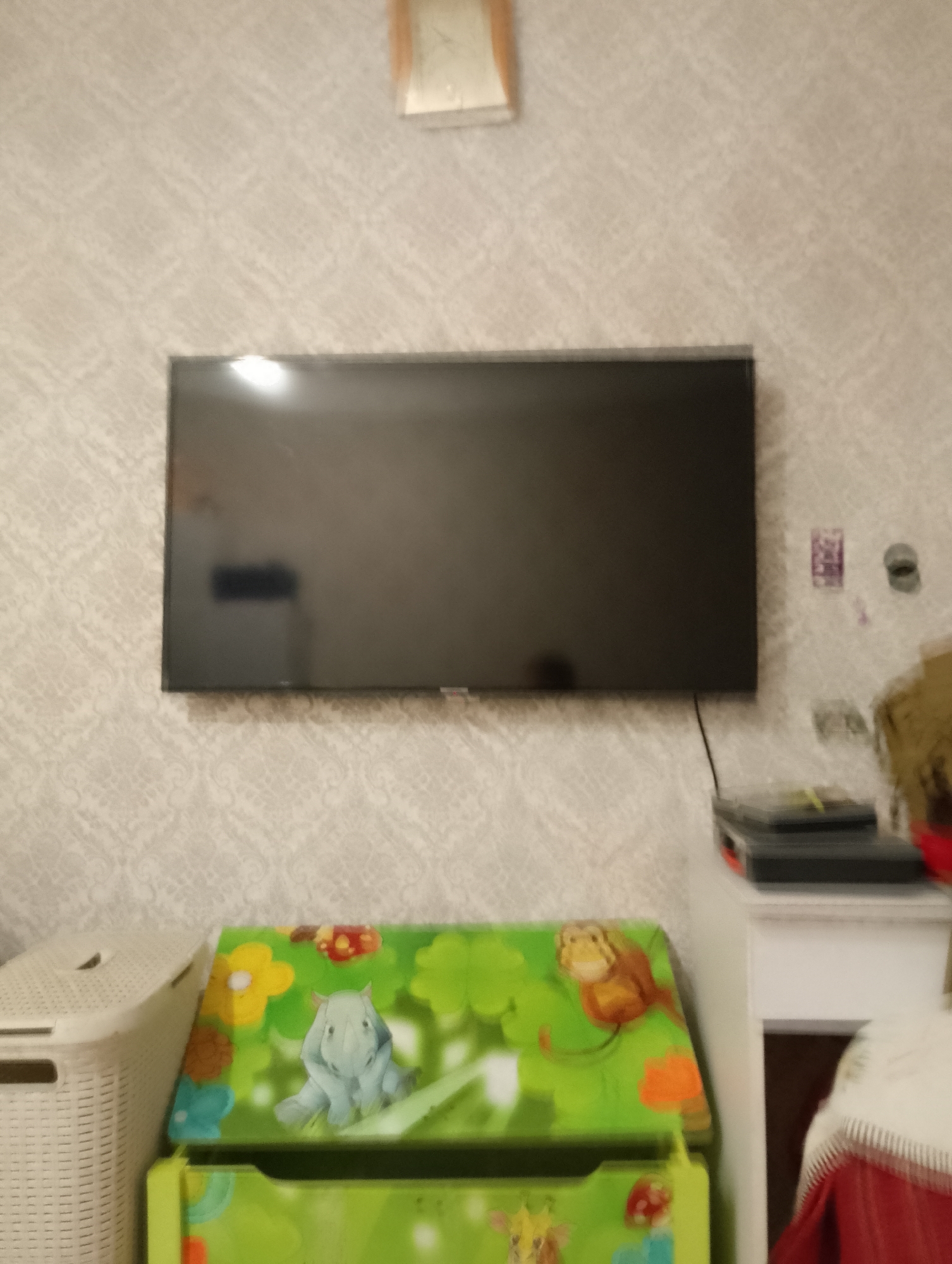 The image shows a television mounted on a wall with a patterned wallpaper. Below the television, there is a toy chest with cartoon animal figures and flowers. To the left of the toy chest, there is a white woven basket. To the right of the toy chest, there is a white cabinet. On the cabinet, there are several stacked items, possibly books or boxes. The scene appears to be in a residential setting, likely a child's room or living room. The image does not provide any information that would allow for localization in Kyiv, Ukraine.