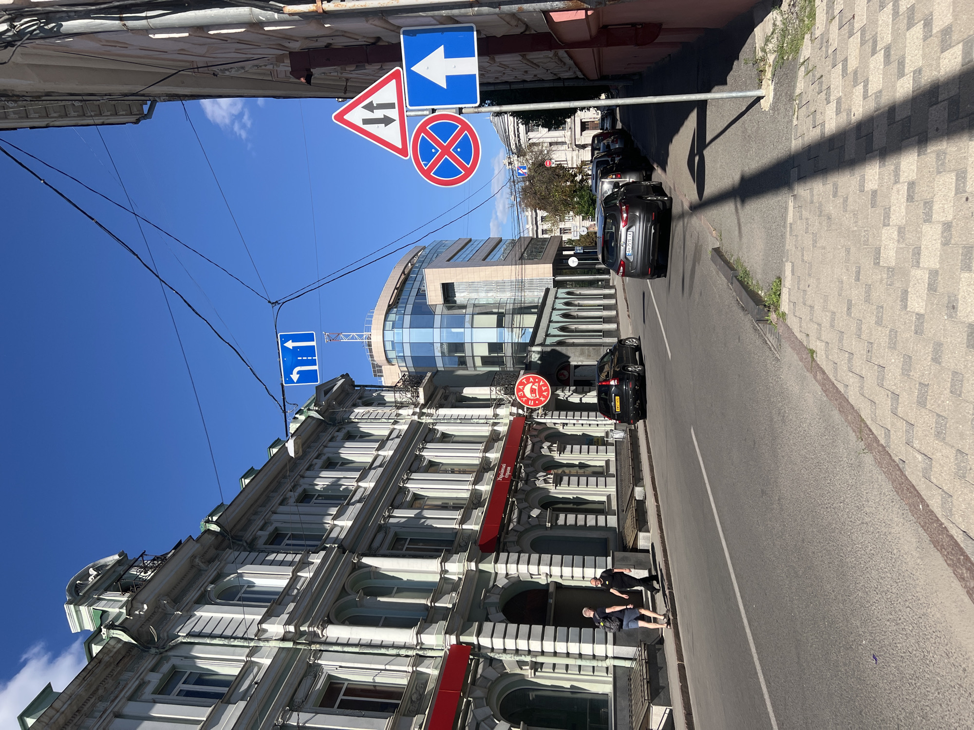 The image shows a street scene in Kharkiv, Ukraine, on a sunny day. A light grey building with ornate architectural details and red awnings is on the left side of the street. In front of this building, two men are walking on the sidewalk. One man, wearing a black t-shirt and pants, is walking towards the right, while another man, wearing a backpack and blue shorts, is walking towards the left. A black SUV is parked on the right side of the street, and several other cars are parked further down. Several traffic signs are visible: a blue sign with a white arrow pointing upwards, a red and white triangular sign with two black arrows pointing downwards, and a circular red sign with a blue circle and a red cross, indicating no stopping or parking. Further down the street, a modern glass-fronted building stands out. The sky is clear blue with a few wispy clouds. The street is paved with asphalt, and the sidewalk is made of interlocking stone tiles.