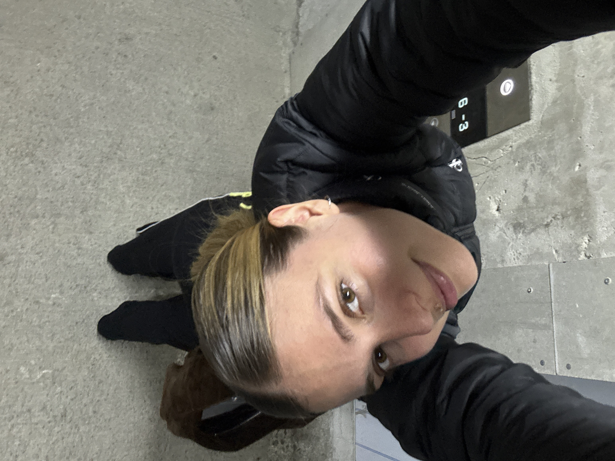 A woman is standing in an elevator lobby in an unknown city. She is wearing a black puffer jacket and black socks, and her hair is pulled back into a ponytail. She is looking up at the camera, smiling slightly. The elevator button panel indicates floors 6-3. The floor and walls are made of concrete.