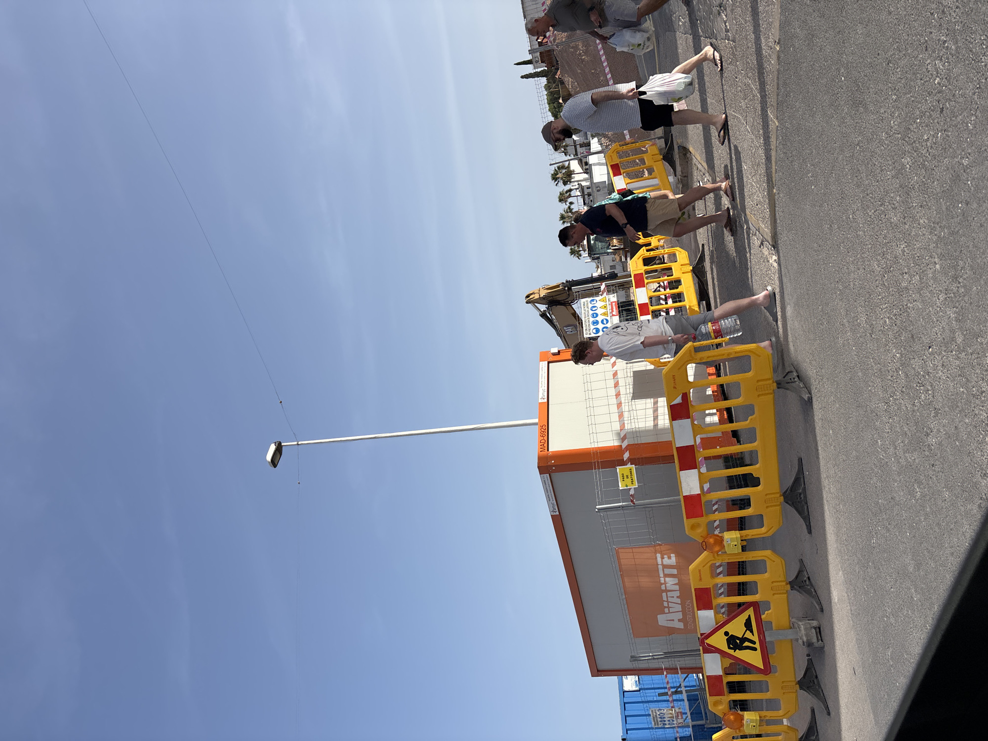 The image shows a group of four men walking across a paved area, likely a street or walkway, in Urbanización Villa Martín, Spain. The area appears to be under construction, with temporary yellow and orange barriers set up to divert pedestrian traffic. A construction trailer, labeled "Avante Construcción," is visible to the left, as is a yellow excavator in the background. The men are dressed in casual summer clothing, and two of them are carrying shopping bags. The sky is clear and blue.