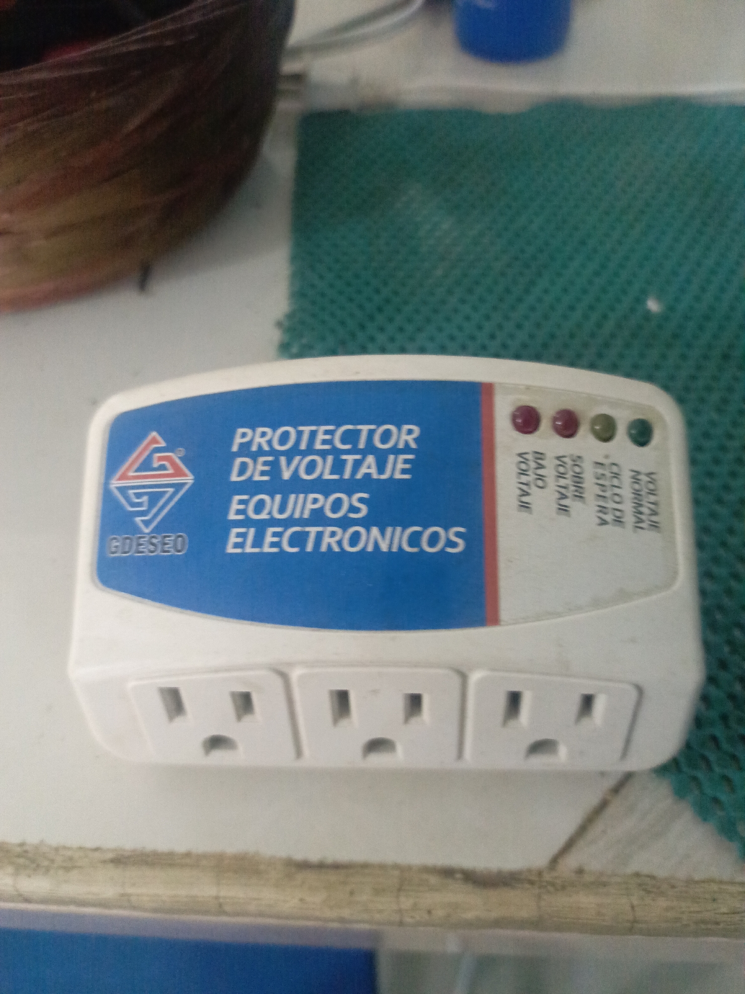 The image shows a GDESEO brand surge protector with three grounded outlets, sitting on a white surface. The protector has a blue label reading "PROTECTOR DE VOLTAJE EQUIPOS ELECTRONICOS" with the GDESEO logo. There are four indicator lights labeled (from left to right) "BAJO VOLTAJE," "SOBRE VOLTAJE," "CICLO DE ESPERA," and "VOLTAJE NORMAL." The lights are red, red, green, and green. A green textured surface is visible behind the protector. The surface the protector is on appears to be slightly dirty. The location information is unknown.