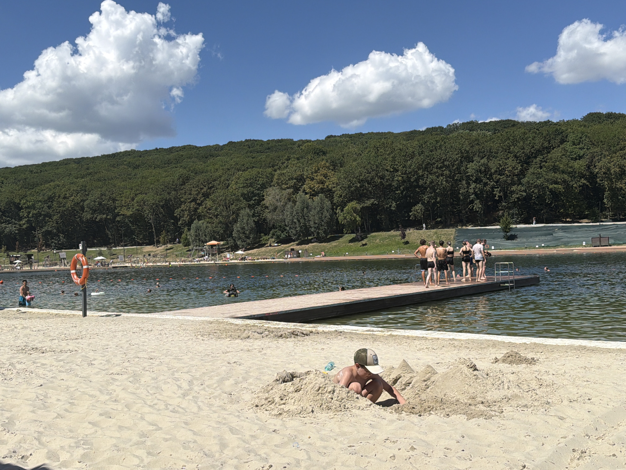 A sunny day at a beach resort in Stavropol, Russia, features a sandy shore in the foreground with a young boy buried up to his chest in the sand, wearing a baseball cap. In the background, a wooden pier extends into a large body of water where several people are swimming and wading. Along the shore, there are lounge chairs, umbrellas, and a lifeguard station. A dense forest covers the hillside overlooking the resort. The sky is blue with fluffy white clouds.