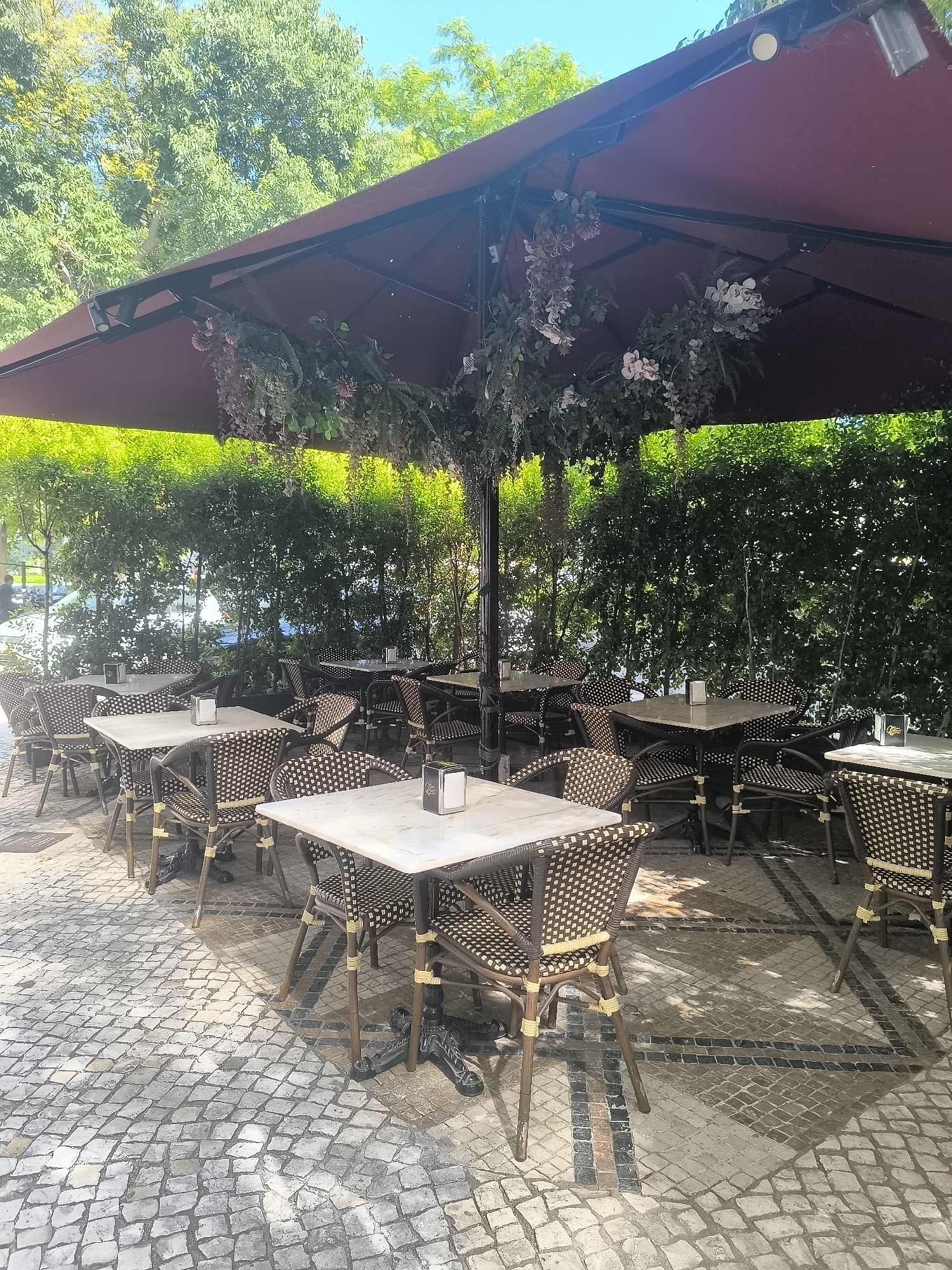 This image shows an outdoor seating area of a cafe or restaurant. Several square marble-topped tables are set with wicker chairs. The tables have dark, ornate metal bases. Each table has a small, silver napkin dispenser. The seating area is covered by a large, dark red umbrella decorated with artificial flowers and greenery. Behind the seating area is a dense hedge of green foliage. The ground is paved with cobblestones. In the background, through the hedge, a street with parked cars and trees is visible under a bright blue sky. The image appears to be taken during the day with ample sunlight casting shadows on the ground. The location is identified as Setúbal, Portugal.