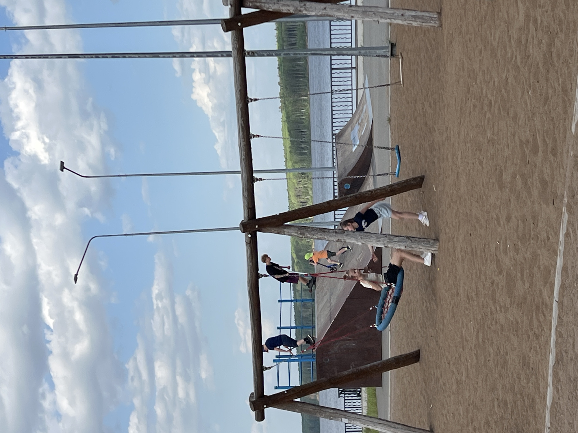 The image shows a playground near the Kama River in Chaikovsky, Russia. There are several children playing on the equipment, including swings and a climbing structure. A group of children is visible on the jungle gym, and one child is swinging in a round swing. The playground is on a sandy surface. In the background, the Kama River is visible, bordered by trees and a metal fence. There are light poles lining the area. The sky is blue with scattered clouds.