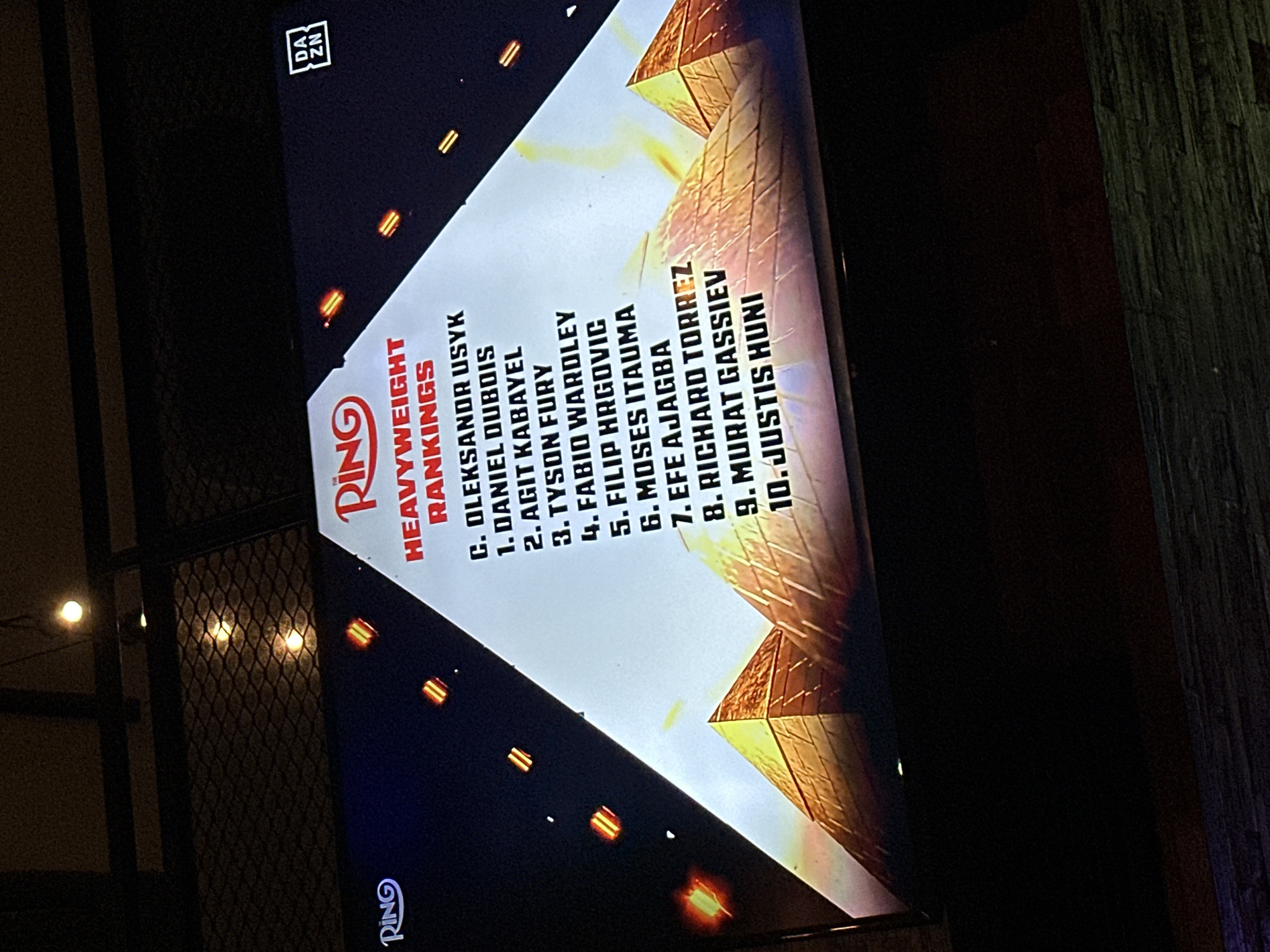 The image shows a television screen displaying "The Ring Heavyweight Rankings." The rankings list ten boxers, starting with "C. OLEKSANDR USYK" and ending with "10. JUSTIS HUNI." The DAZN logo is visible in the upper right corner. The photograph was taken indoors. The fence/grill outside is indicative of a setting in the vicinity of North Kuta, Indonesia.