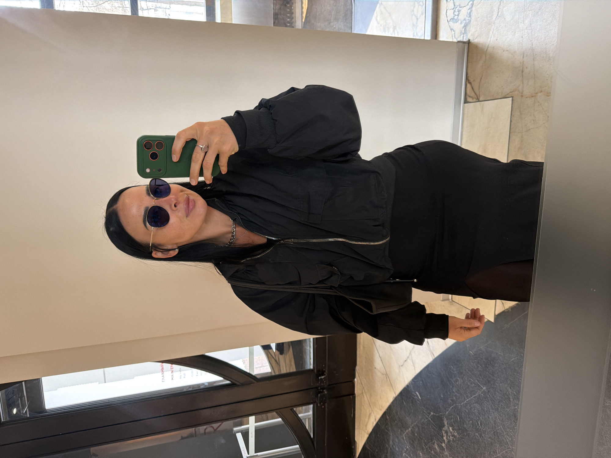 A woman with dark hair and sunglasses takes a selfie in a mirror. She is wearing a black dress and a black bomber jacket. Her phone is green. The background appears to be an interior space with large windows and a decorative floor, suggesting a commercial or public building. Based on the text visible through the window (fragments of Russian words and opening hours), it is plausible that the location is in Saint Petersburg, Russia.
