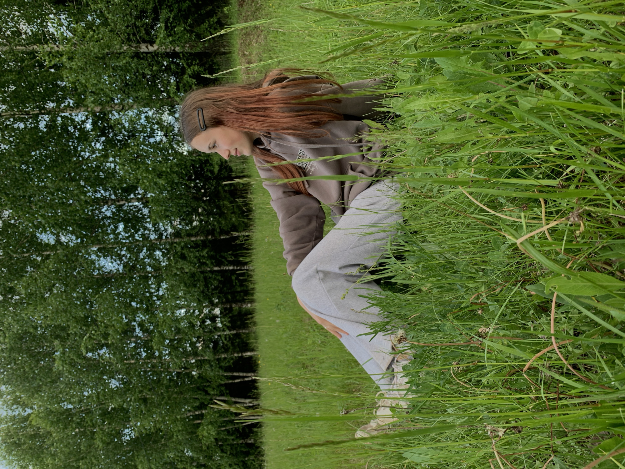 A solitary individual, appearing female, is depicted seated in a natural, overgrown grassy field in Фоки, Russia. The individual, positioned slightly right of the frame's center, has long, reddish-brown hair with a dark, oval-shaped hair clip on the right side. She wears a brown hooded sweatshirt with a white graphic print on the front and light grey sweatpants, complemented by light-colored sneakers. Her posture indicates a relaxed recline, with legs extended and slightly bent, and her left hand resting on her left knee. Her head is turned to the right, and her gaze is directed horizontally into the distance, with a subtle, serene expression. The foreground and midground are dominated by dense, tall green grass and various wild plants, some with visible seed heads. In the background, a thick line of trees, identifiable as birches by their light-colored trunks and abundant green foliage, forms a natural barrier. Patches of light sky are visible through the tree canopy. The scene is illuminated by natural daylight.